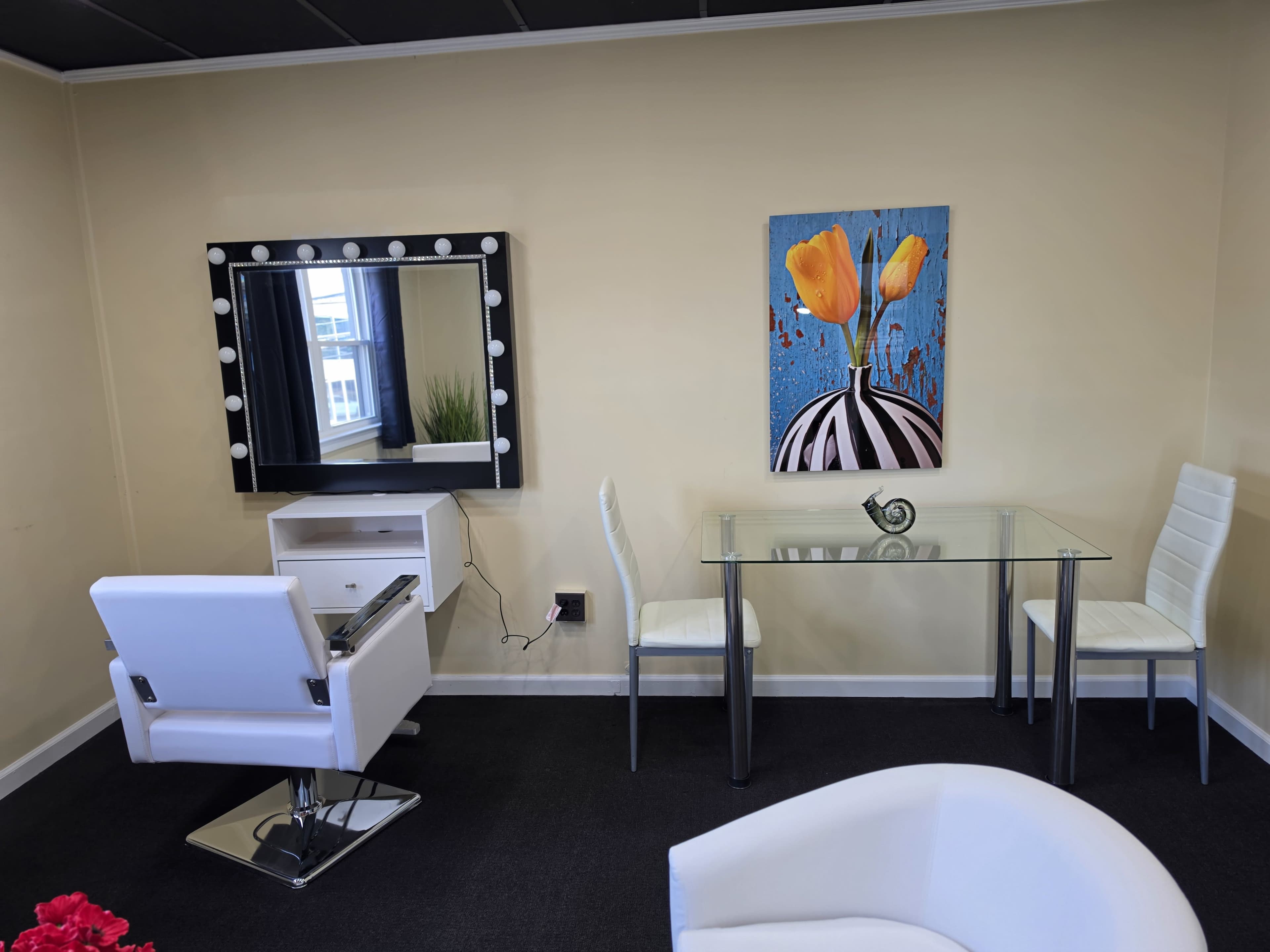 A well-lit salon interior features a makeup mirror with lights, a glass table with two chairs, and a modern styling chair, all set against a beige wall and a colorful flower painting.