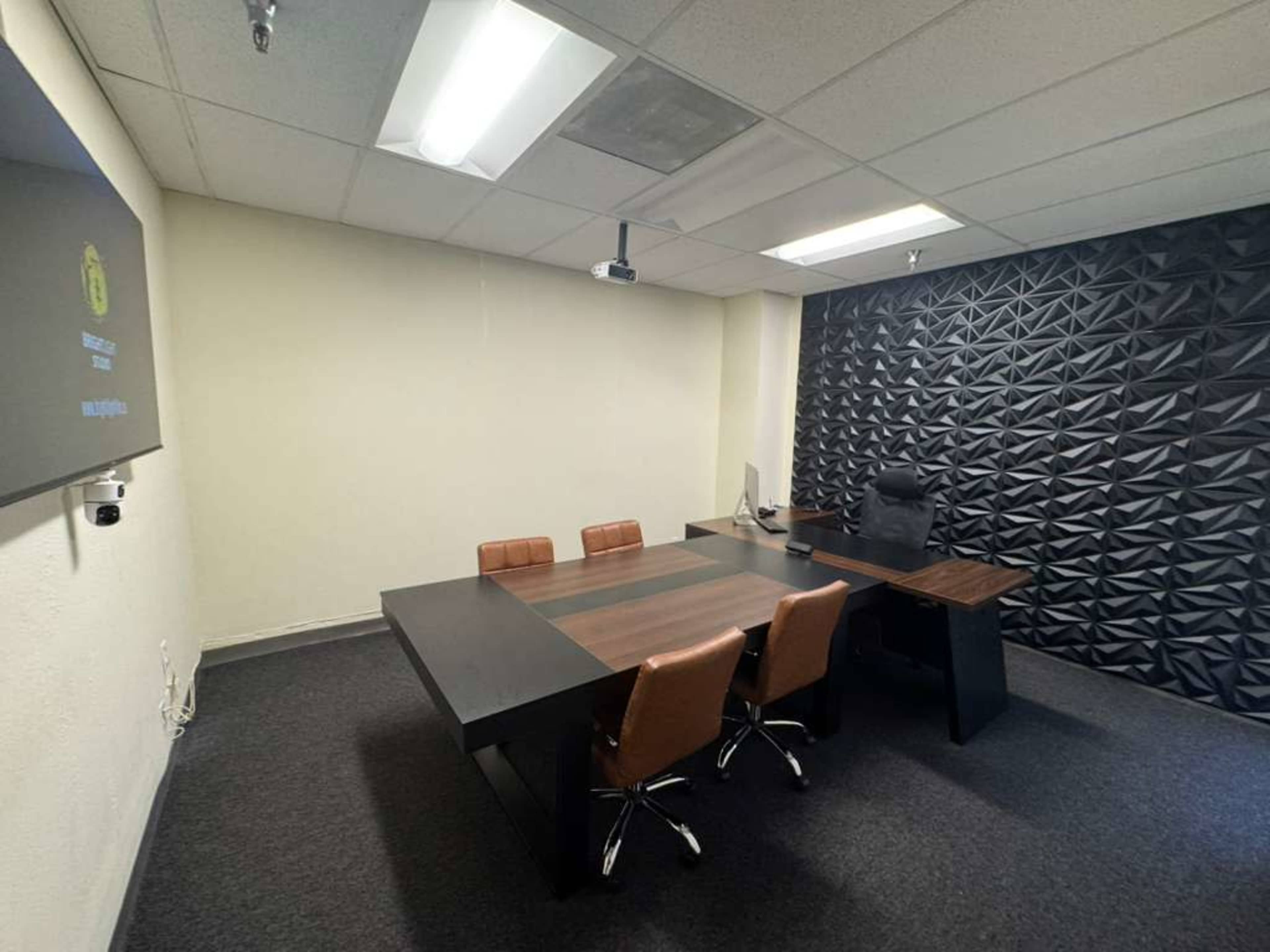 The image shows a modern office with a conference table surrounded by four chairs, a computer workstation, and a textured black wall.