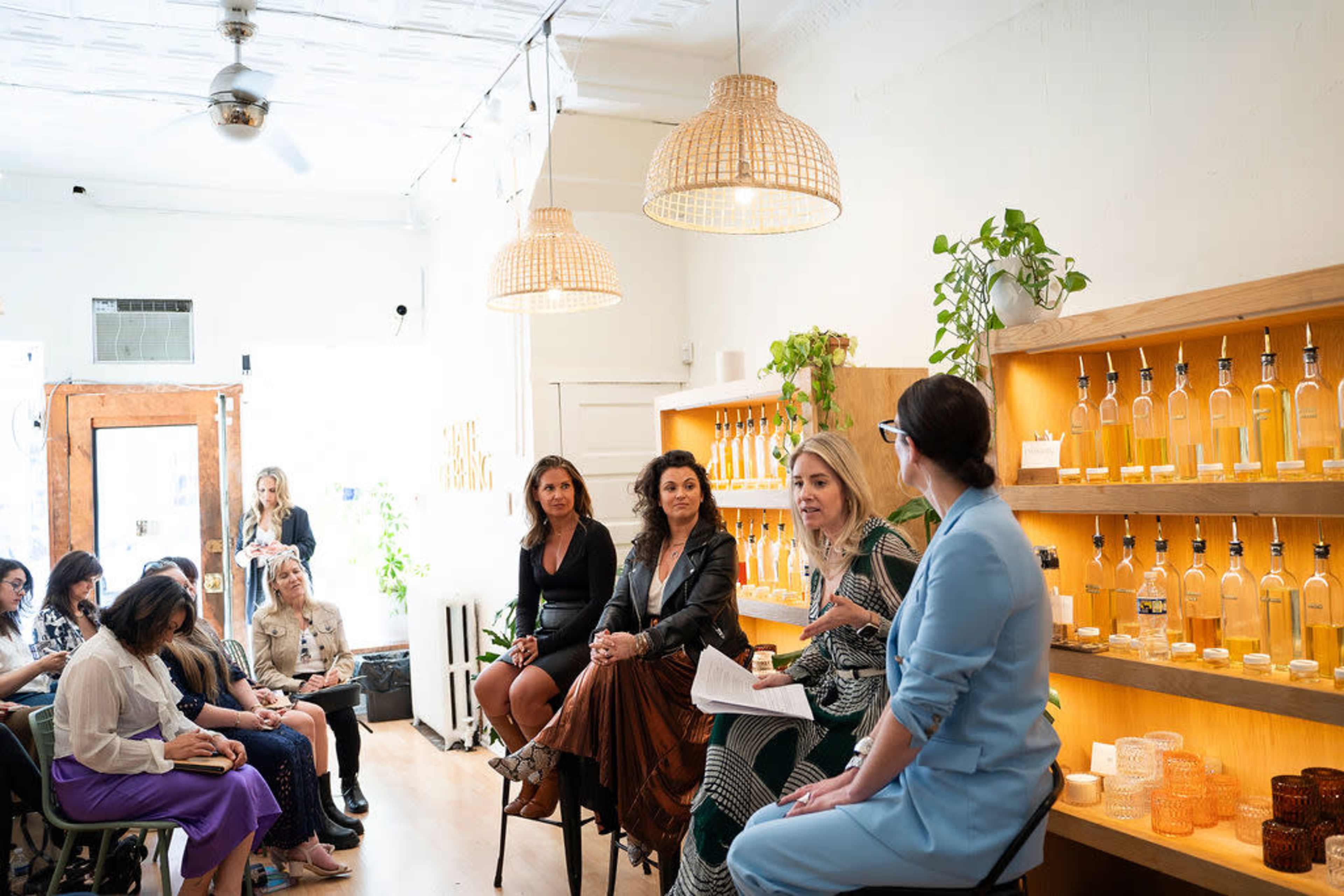 A group of women engage in a discussion in a brightly lit shop with plants and bottles displayed on the shelves, while an audience listens attentively.