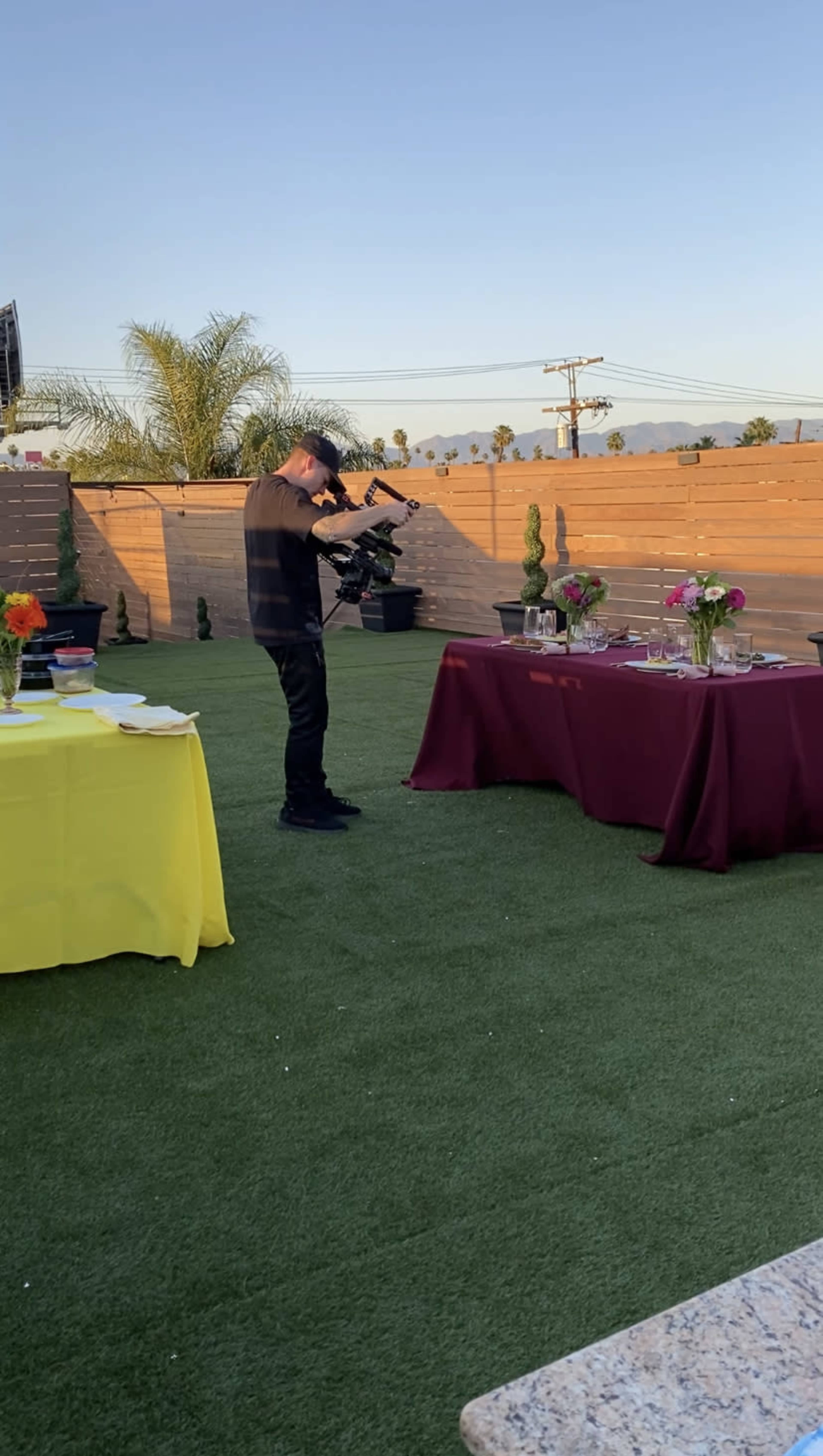 A person is filming a setup with tables covered in yellow and burgundy cloths, adorned with floral centerpieces, in an outdoor space.