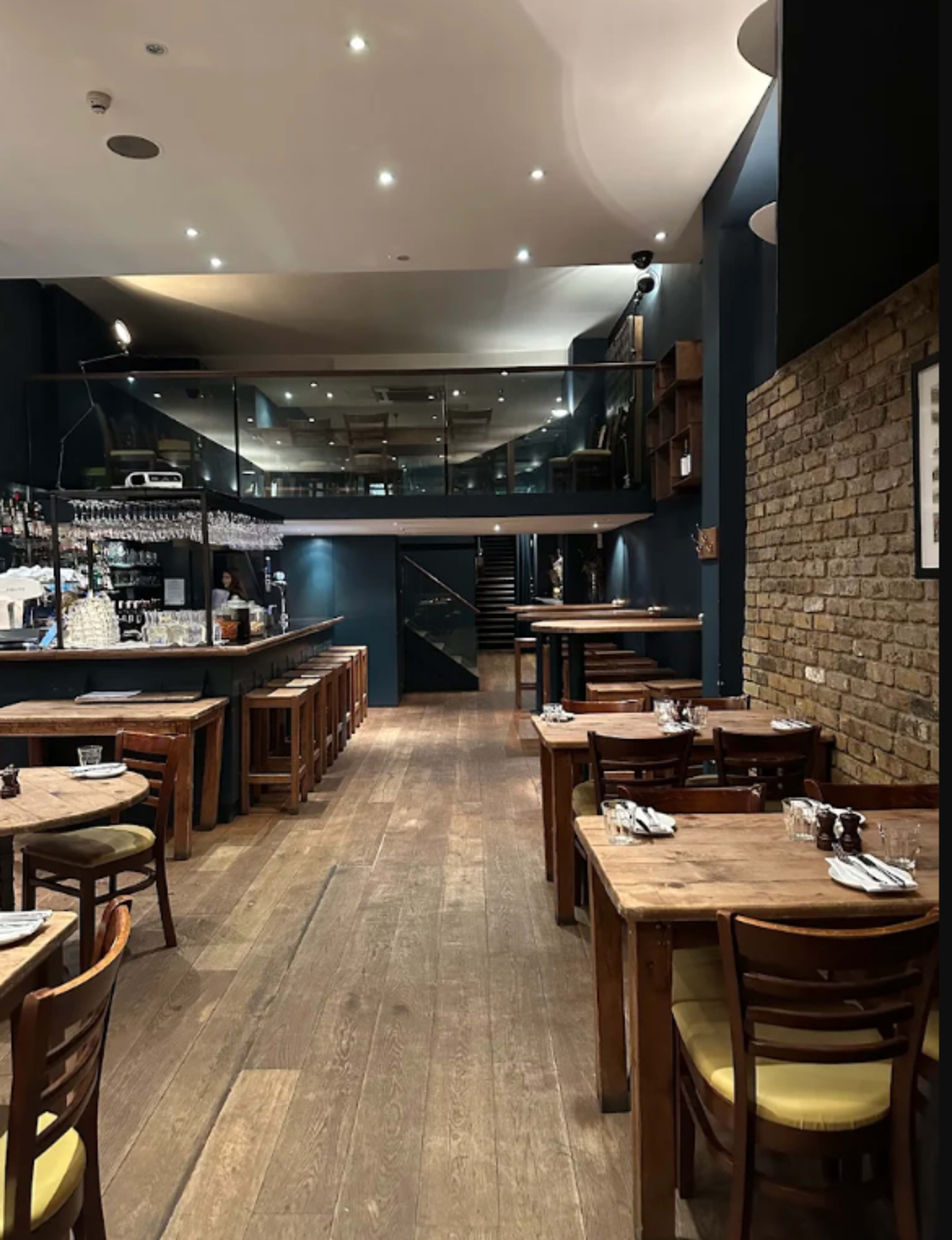 The image shows an interior view of a restaurant featuring wooden tables and chairs arranged on a wooden floor, with a bar area and a staircase visible in the background.
