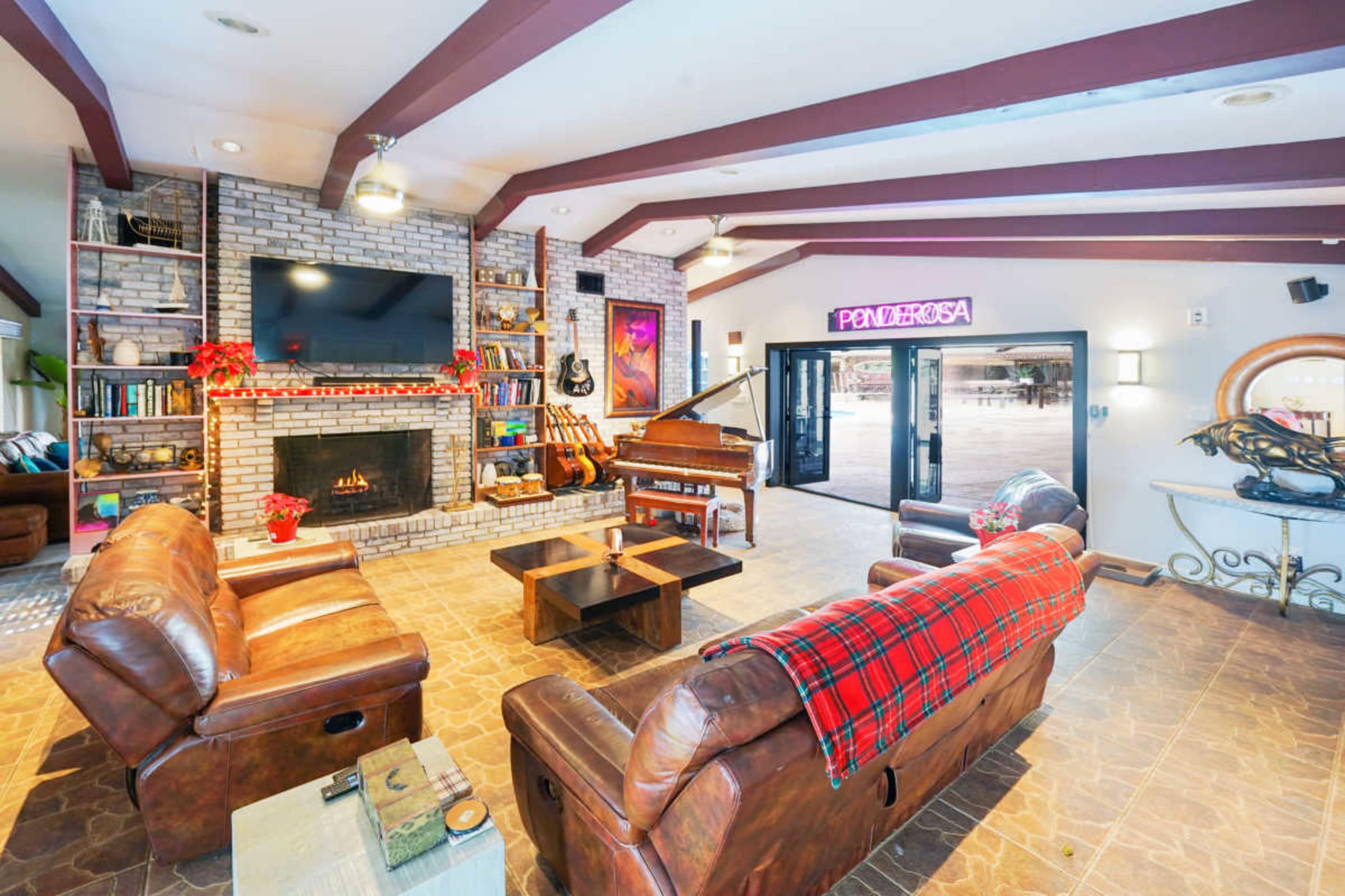 The interior of a living room features leather sofas, a piano, a fireplace, and a large television, with doors leading outside and decorated for the holiday season.