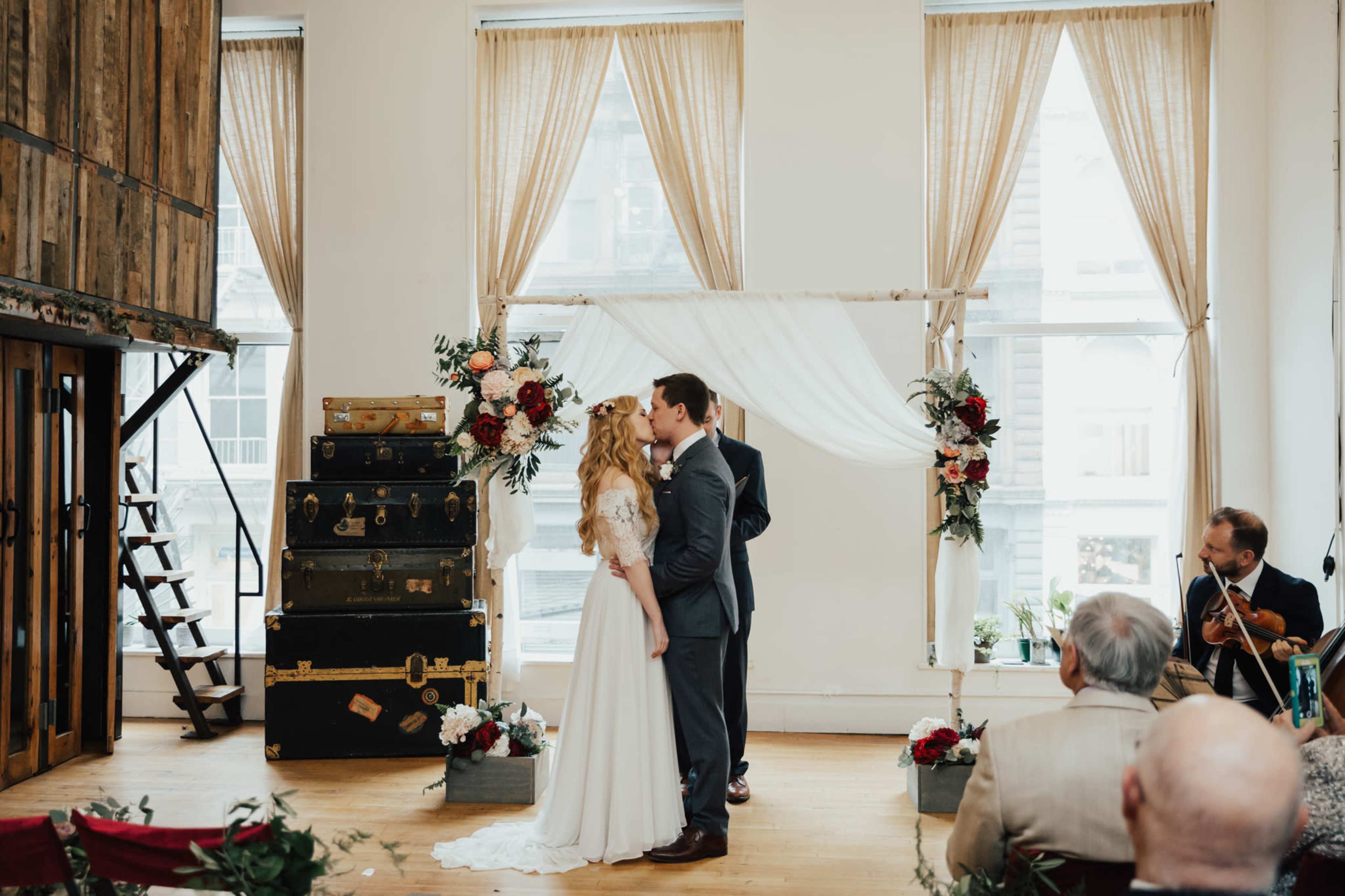 A couple stands under a floral arch, exchanging a kiss during their wedding ceremony in a spacious room filled with natural light.