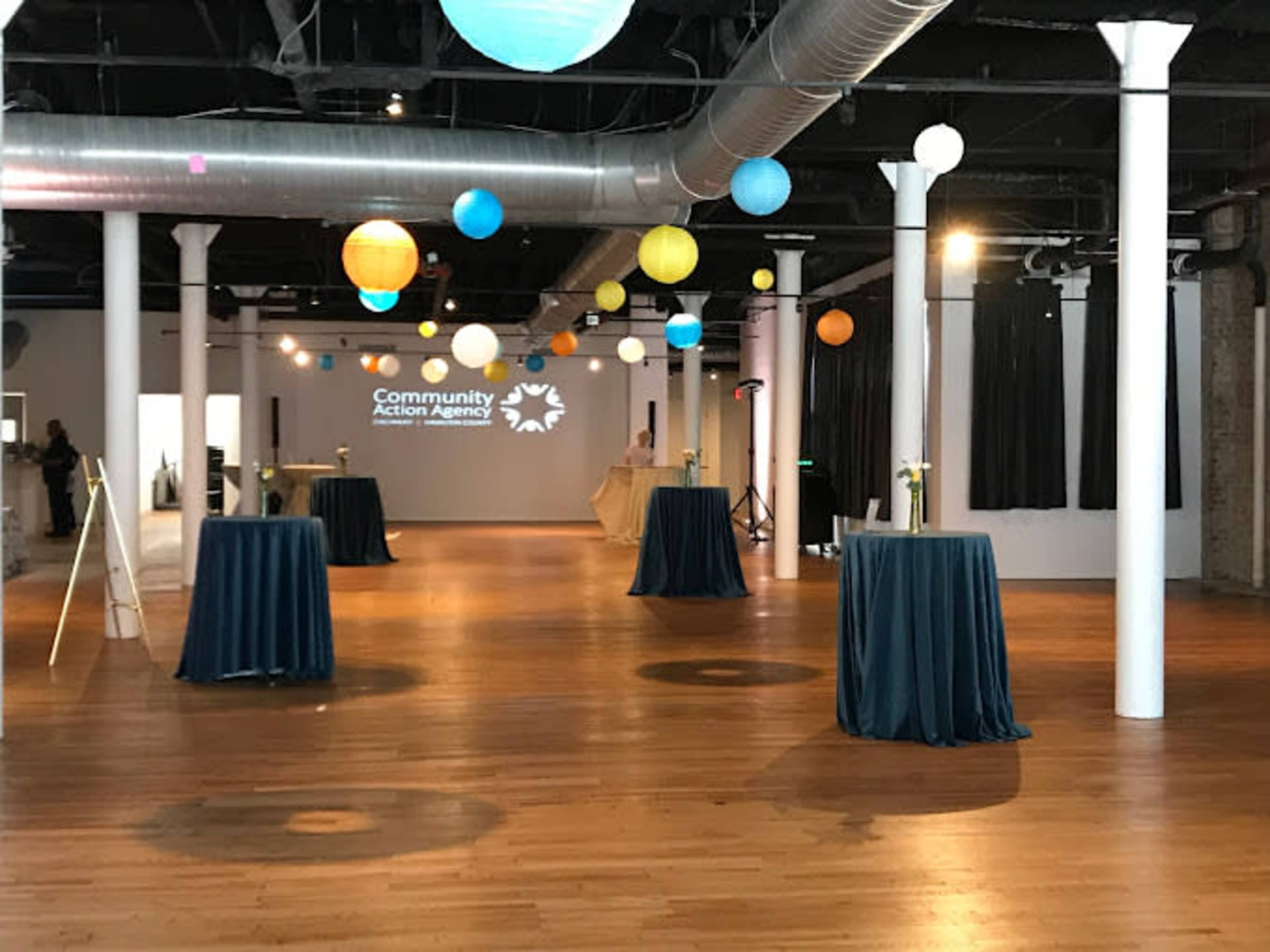 A spacious event venue features tall tables covered in blue cloth, colorful hanging lanterns, and a projection of the Community Action Agency logo on the wall.