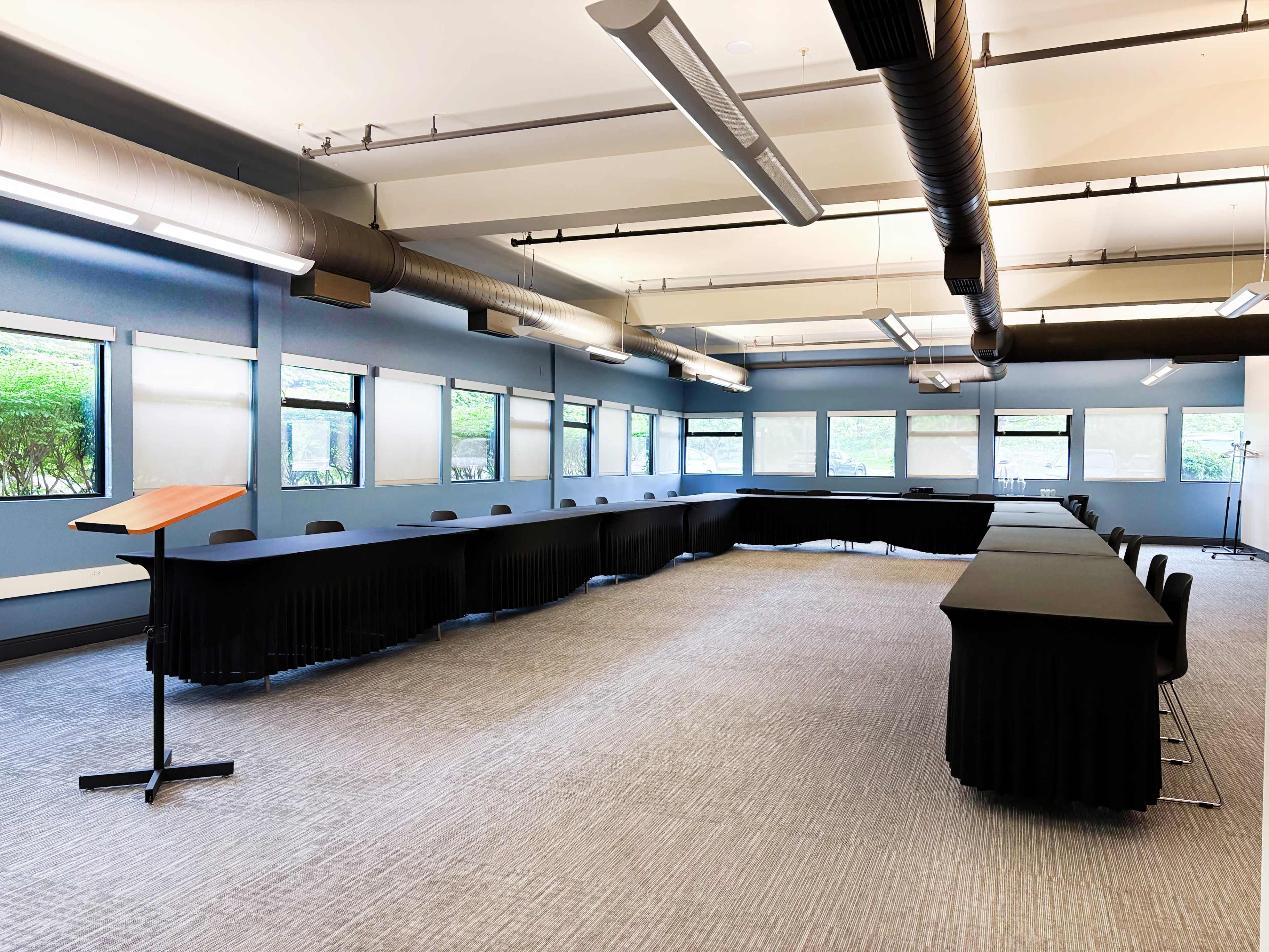 The image shows a spacious conference room with a long, curved table and rows of black chairs, illuminated by overhead lights and natural light from large windows.