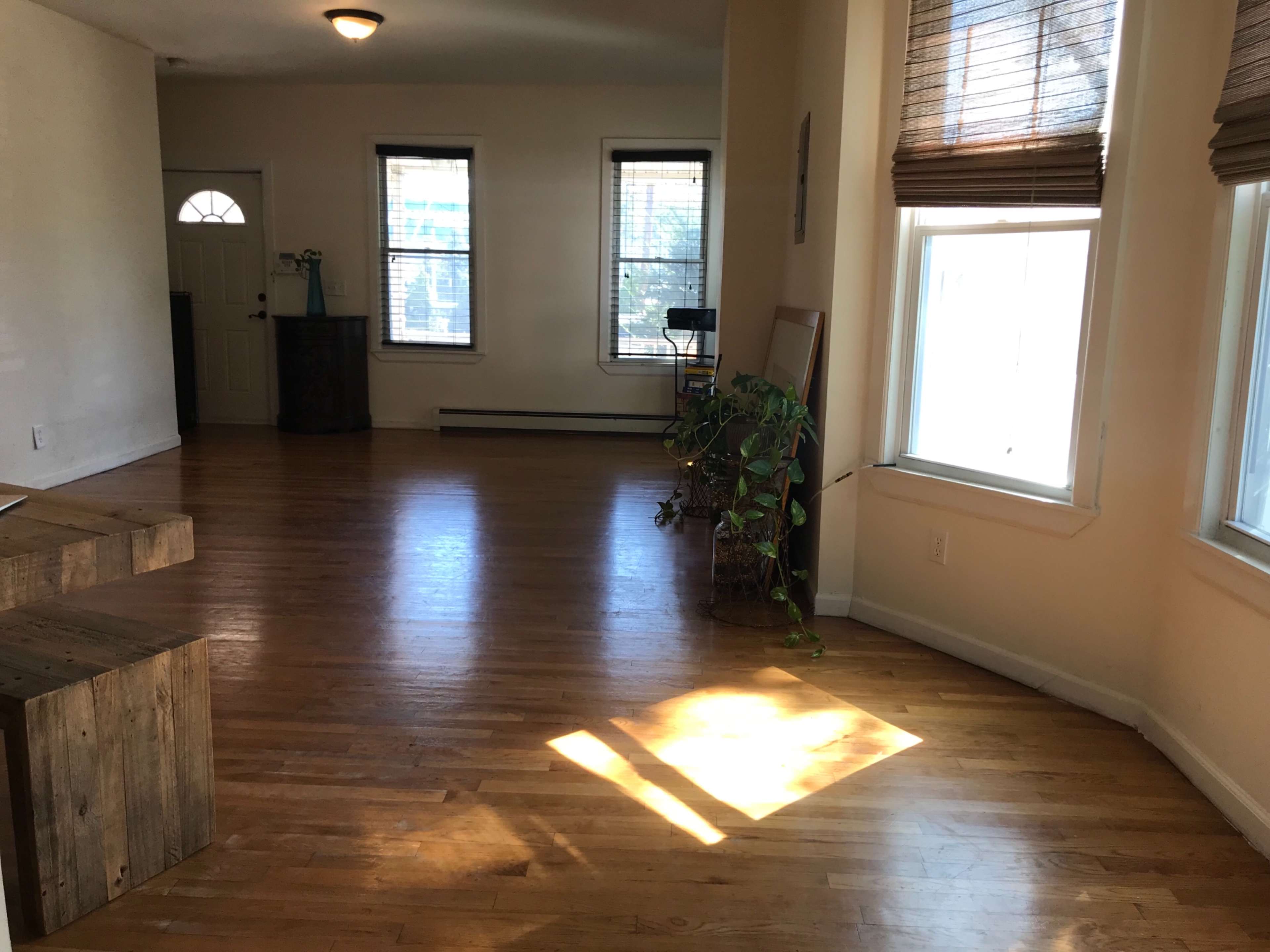 The image shows a spacious room with hardwood floors, two windows letting in natural light, and a wooden bench against one wall.