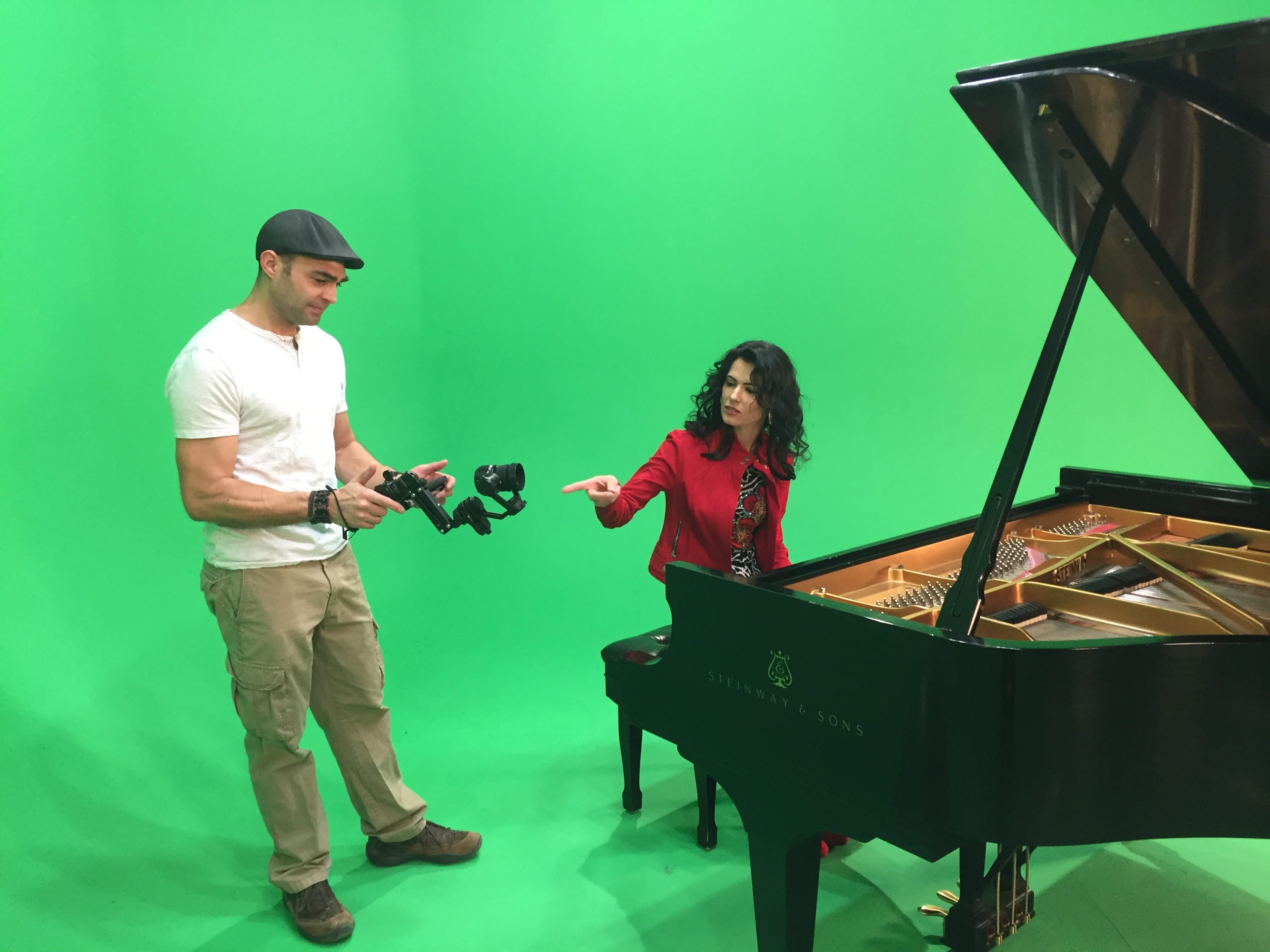 A man holding a camera stabilizer stands next to a woman in a red outfit, who is pointing at a grand piano against a green screen backdrop.