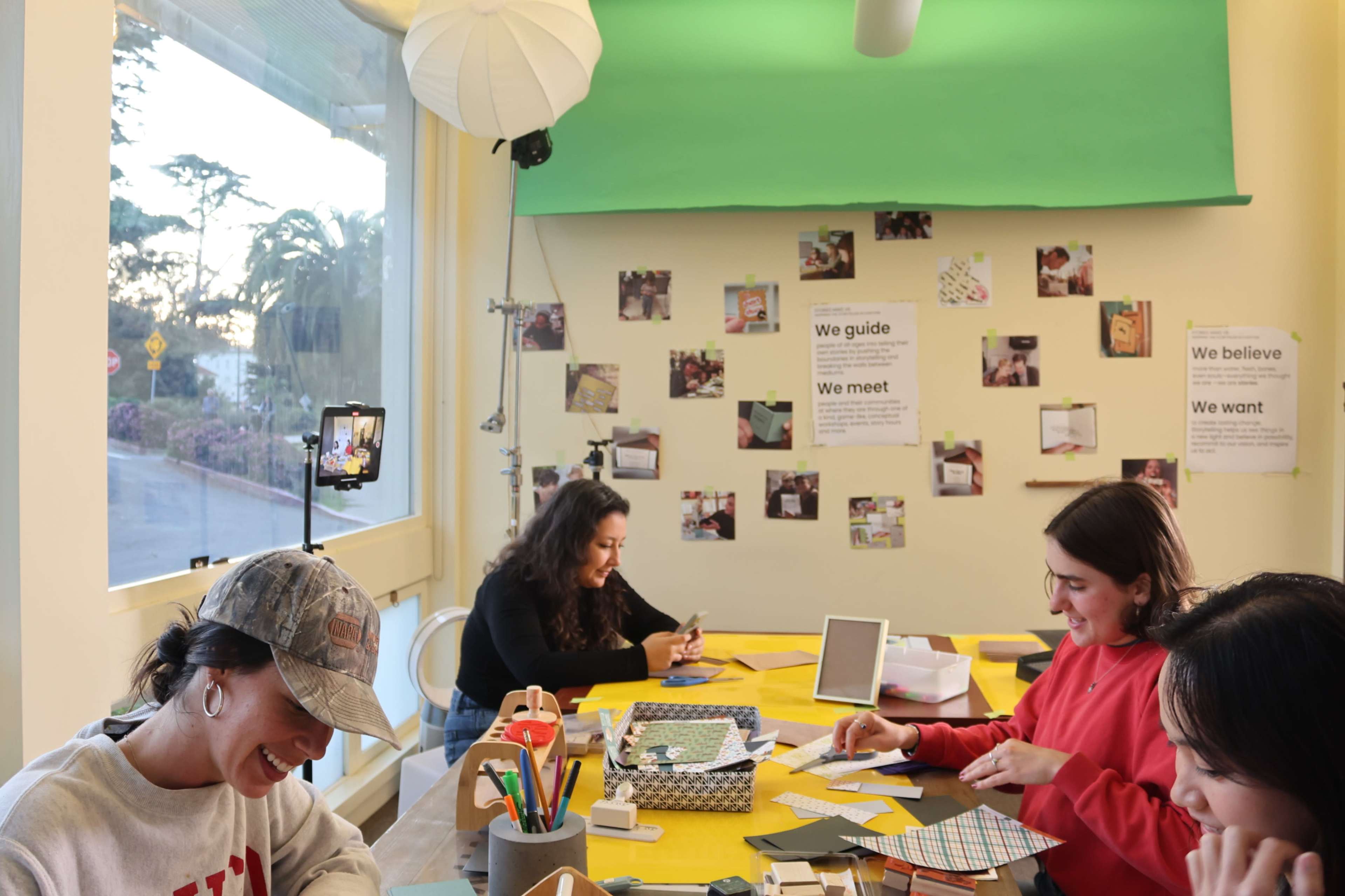 Four people are engaged in a crafting activity at a table adorned with colorful papers and tools, while a wall displays various photos and motivational texts.