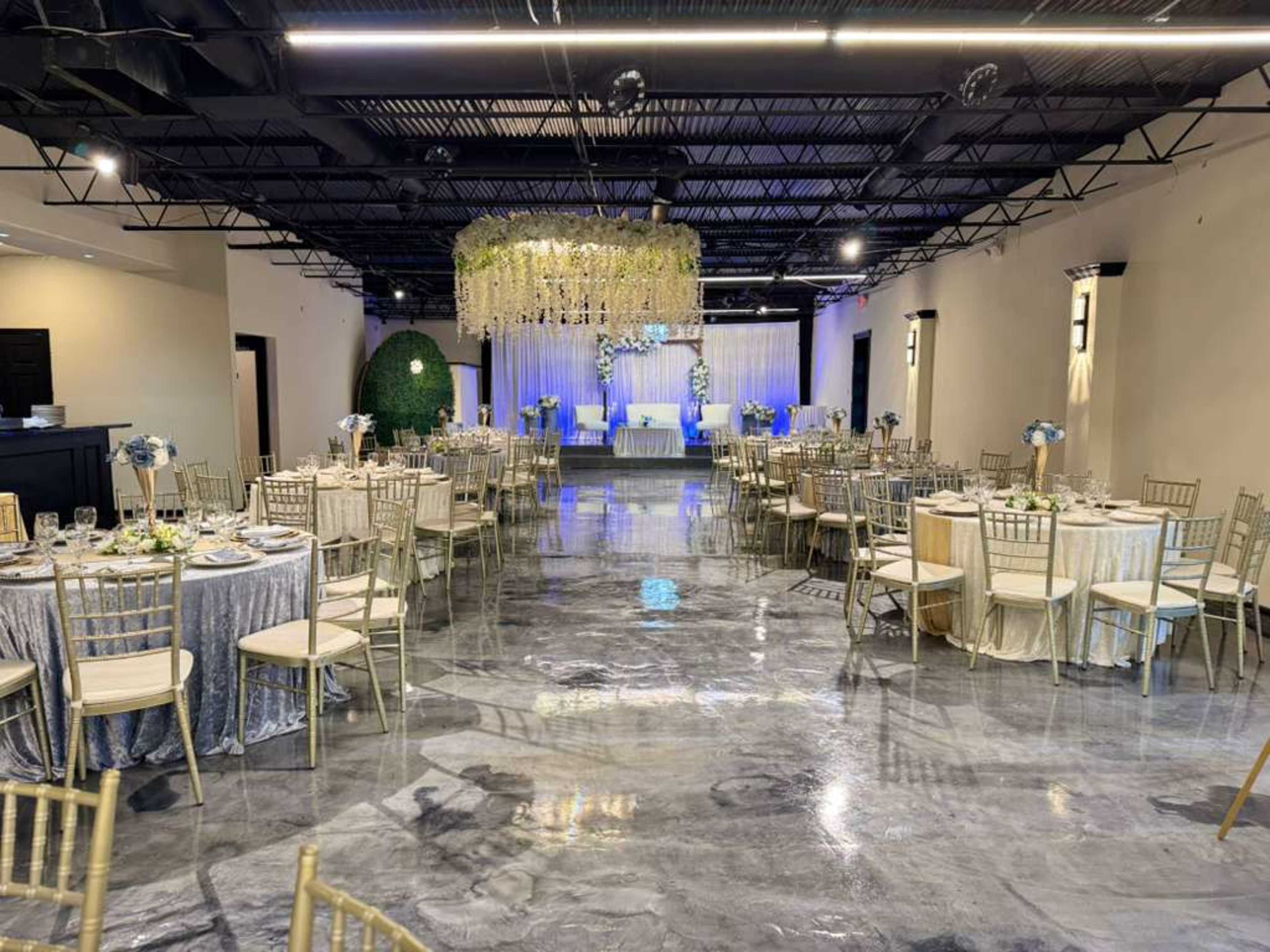 The image shows a spacious event venue with round tables set for dining, decorated with floral centerpieces and a stage at the front, under a ceiling adorned with hanging decorations.