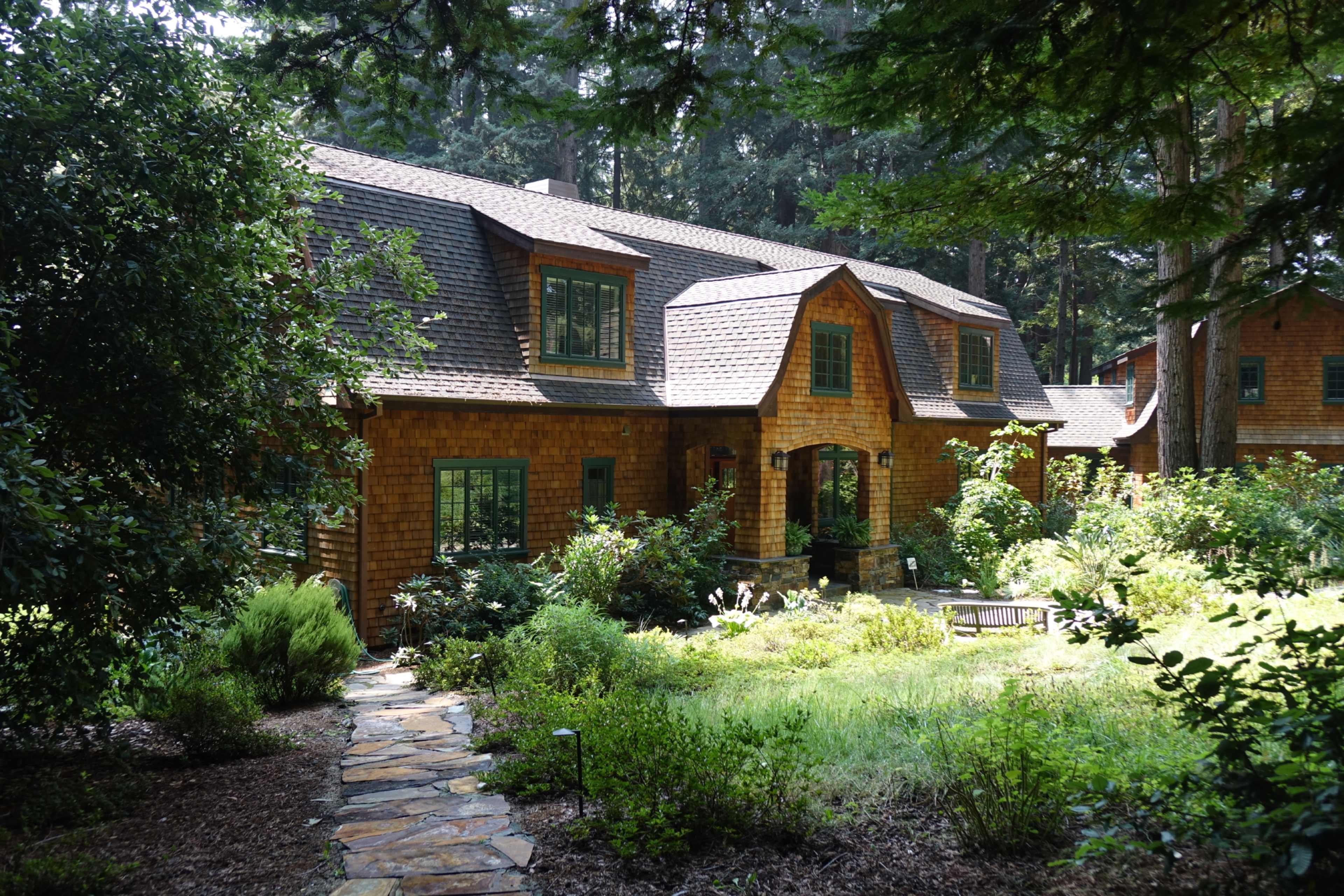 A multilevel wooden house with a shingled exterior is surrounded by dense greenery and a stone pathway.