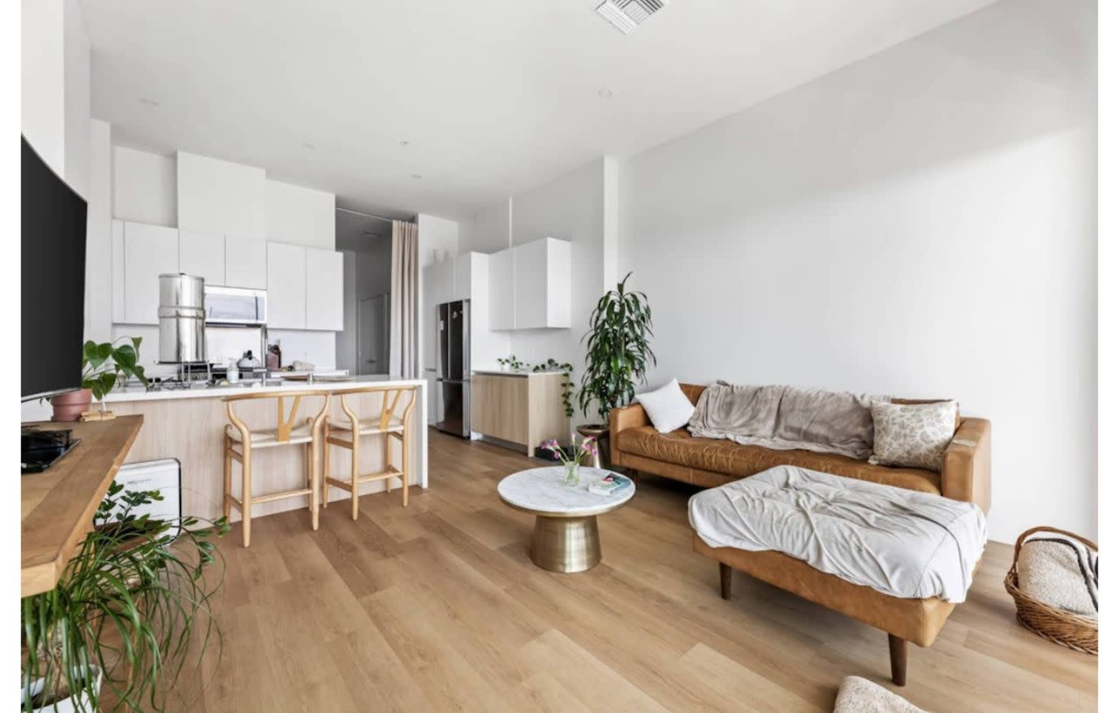 The image shows a modern, open-concept living space featuring a kitchen, a dining area with wooden chairs, and a cozy living room with a tan leather sofa and a round coffee table.