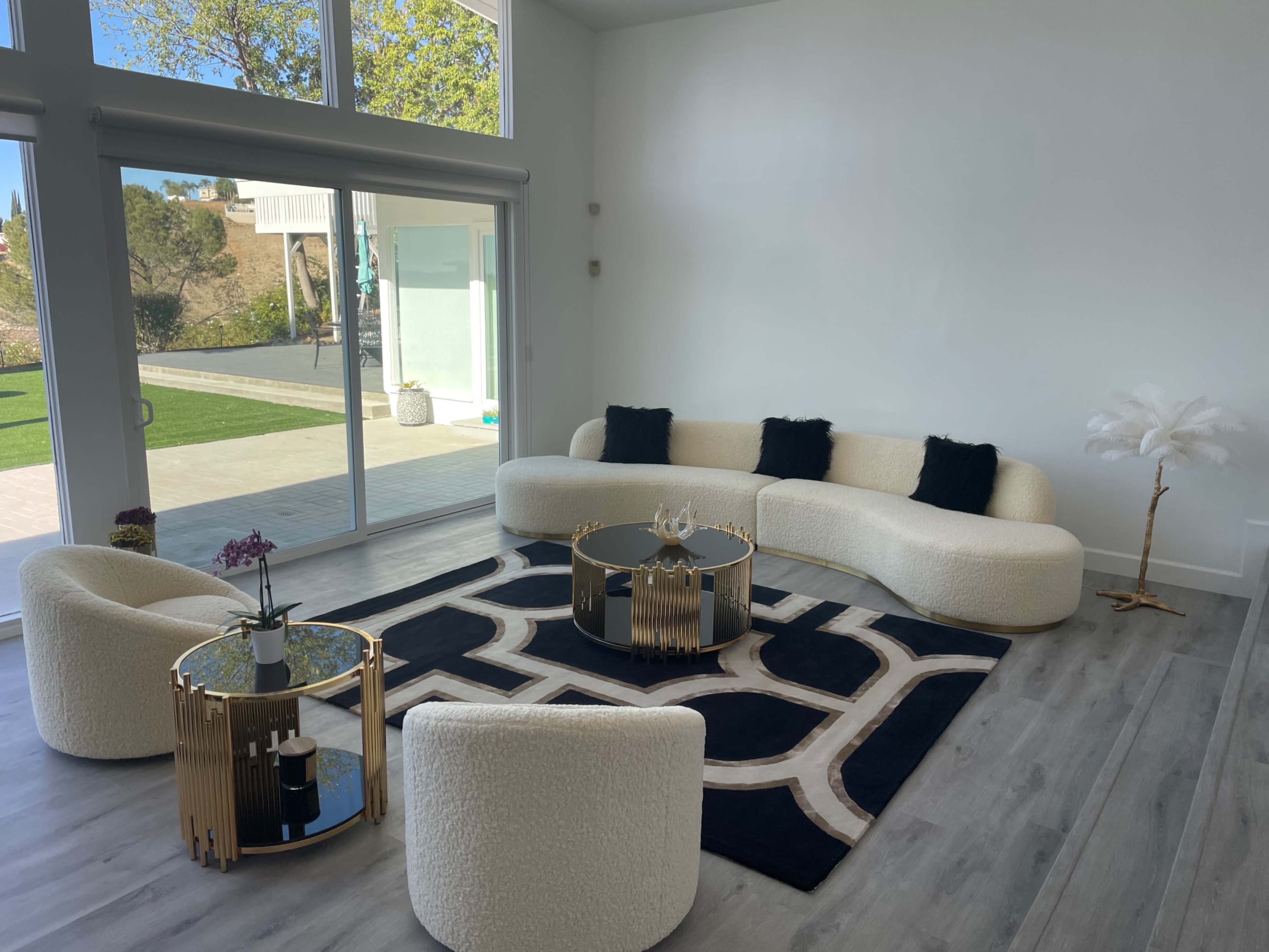 A modern living room features a curved white sofa, two matching chairs, and a geometric rug, arranged around black and gold coffee tables, with large windows overlooking a green outdoor space.