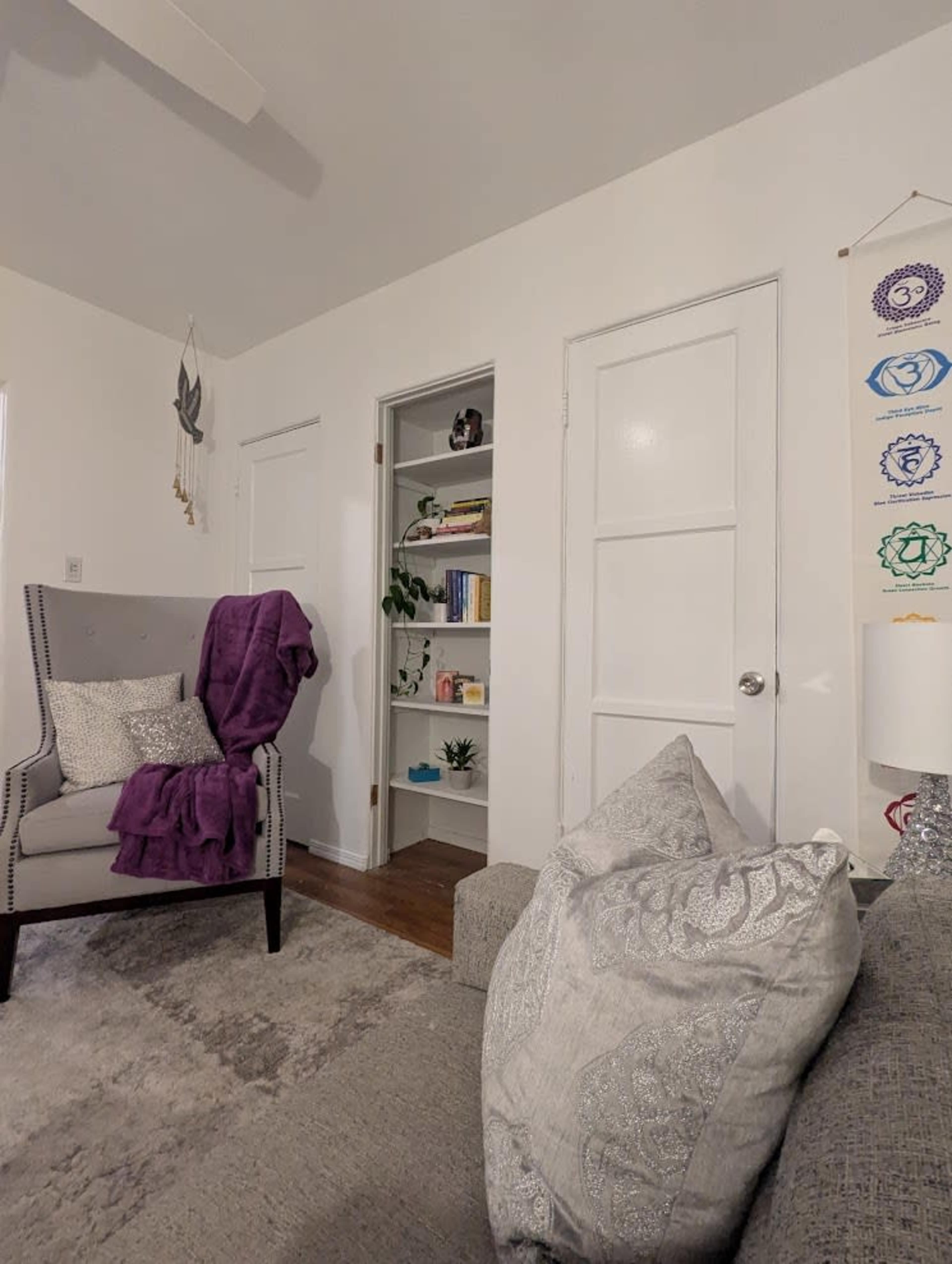 The image shows a cozy living room with a gray armchair draped in a purple throw blanket, a shelf with books and plants in the background, and white walls.