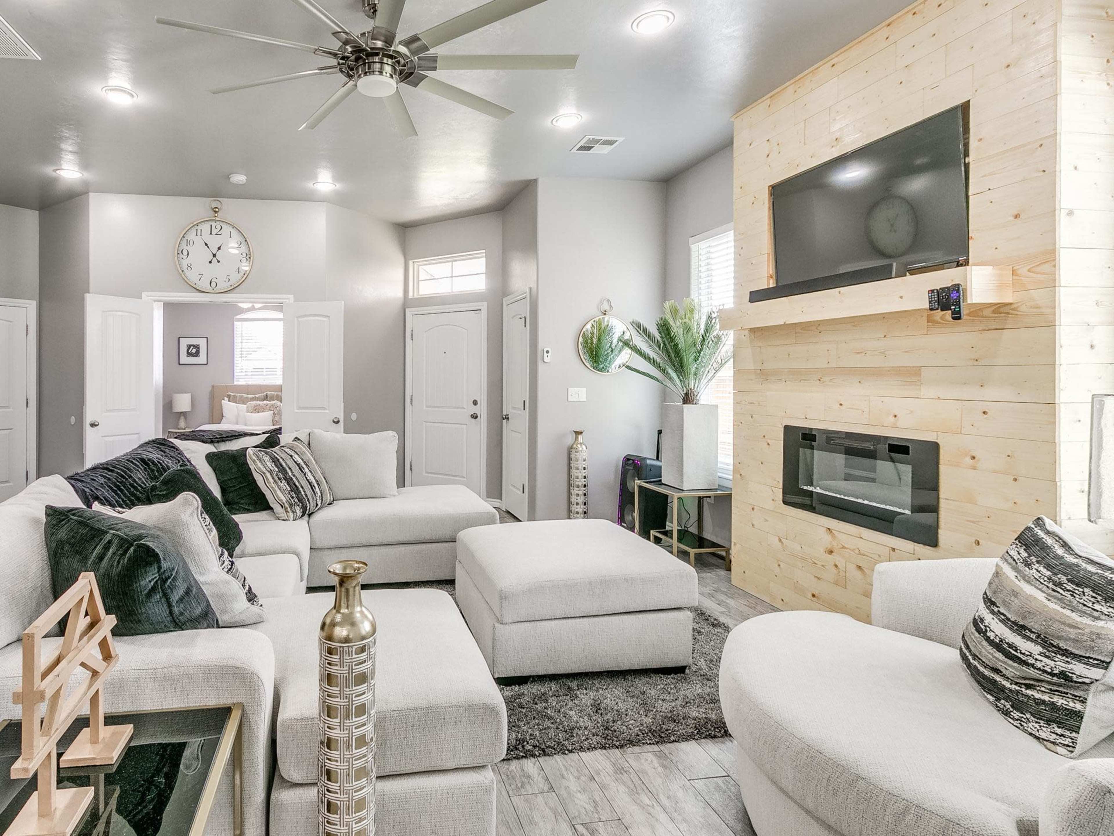 A modern living room features a large sectional sofa, a wooden accent wall with a mounted TV, and a combination of light and dark decor elements.