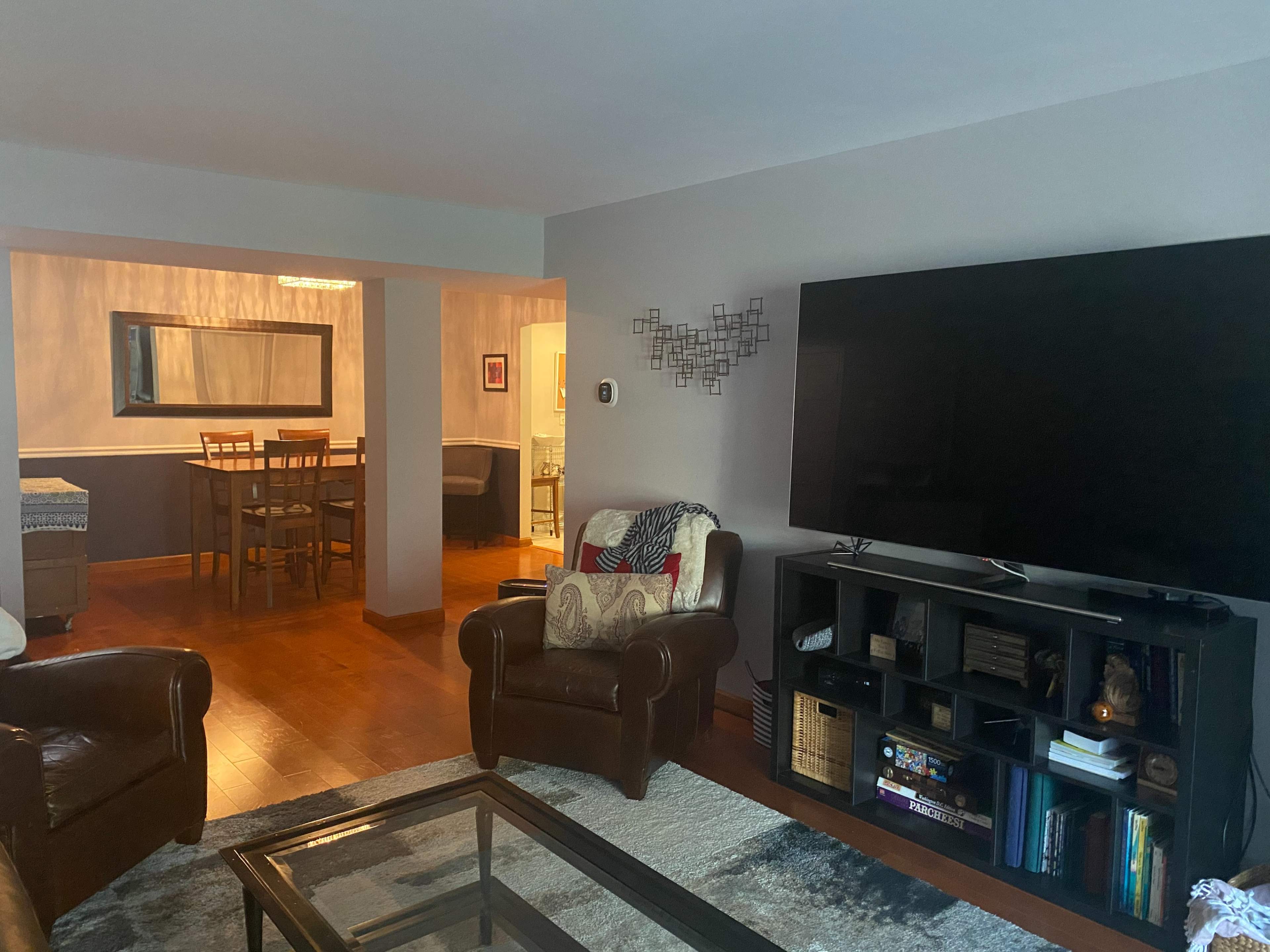 A living room with leather chairs, a glass coffee table, and a large television mounted on a dark shelf, with a dining area visible in the background.