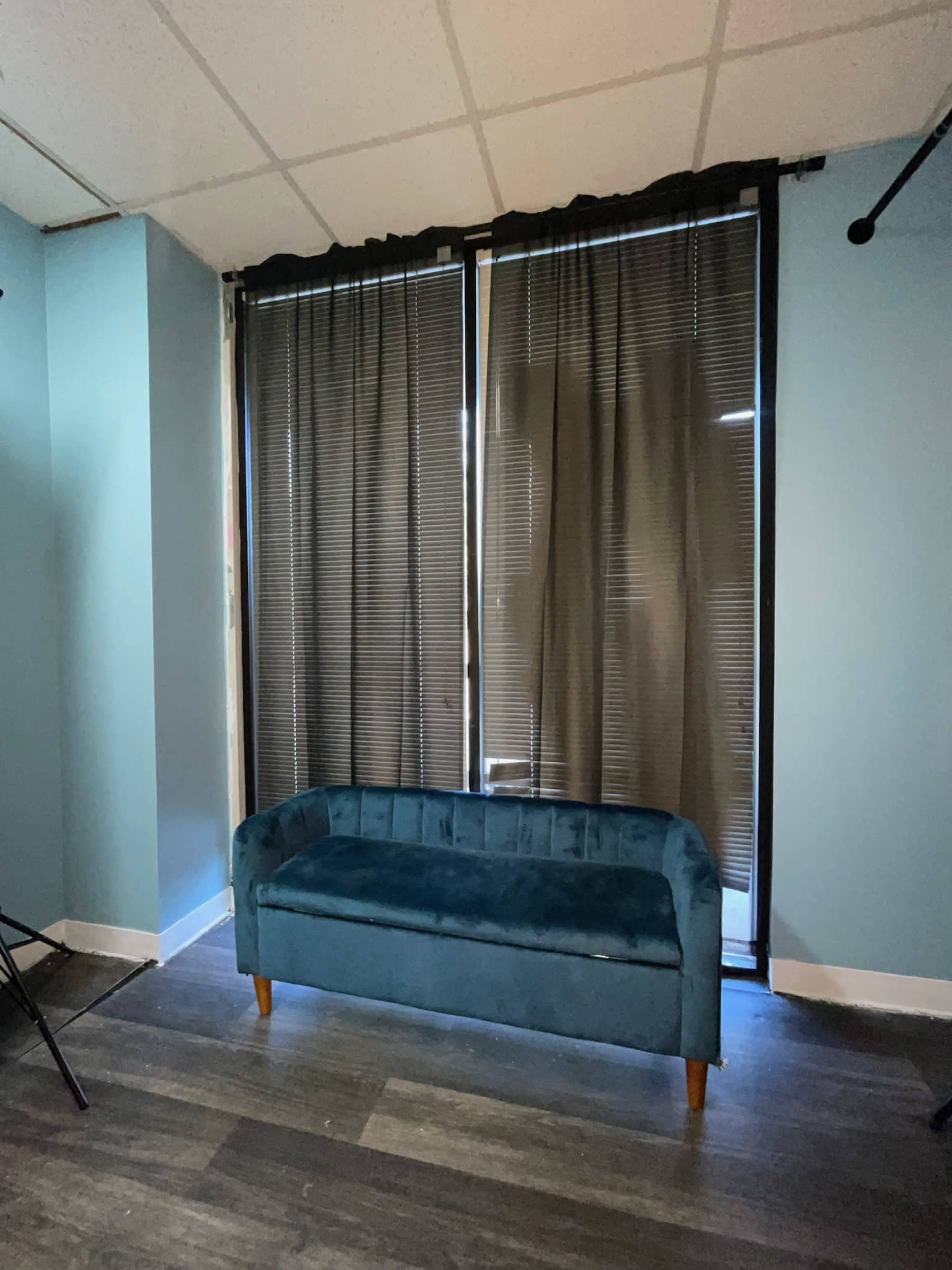The image shows a teal couch placed in front of large black curtains covering a window within a room with light blue walls.