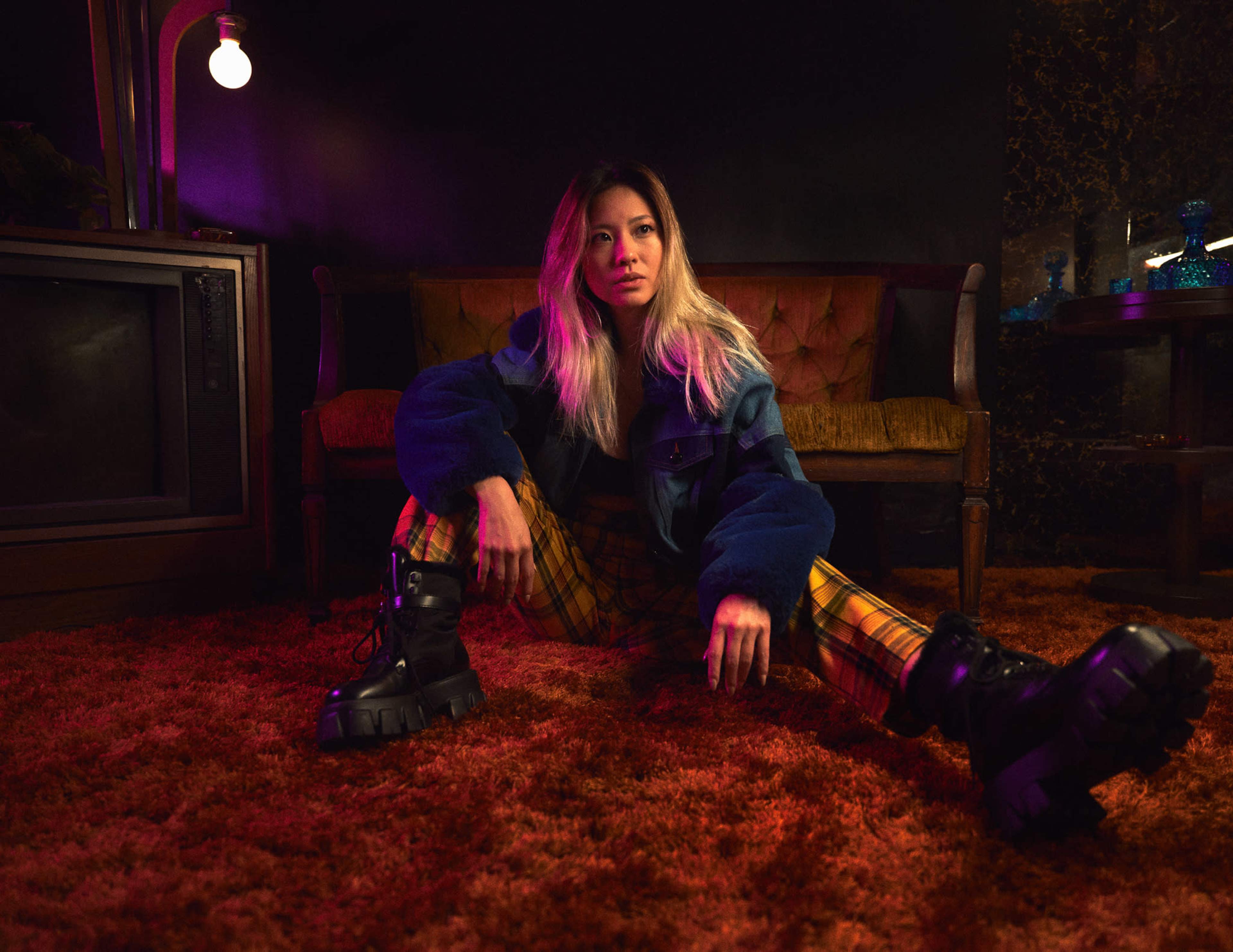 A woman sits on a carpeted floor in front of a vintage couch, wearing a jacket and plaid pants, with an illuminated light bulb overhead.