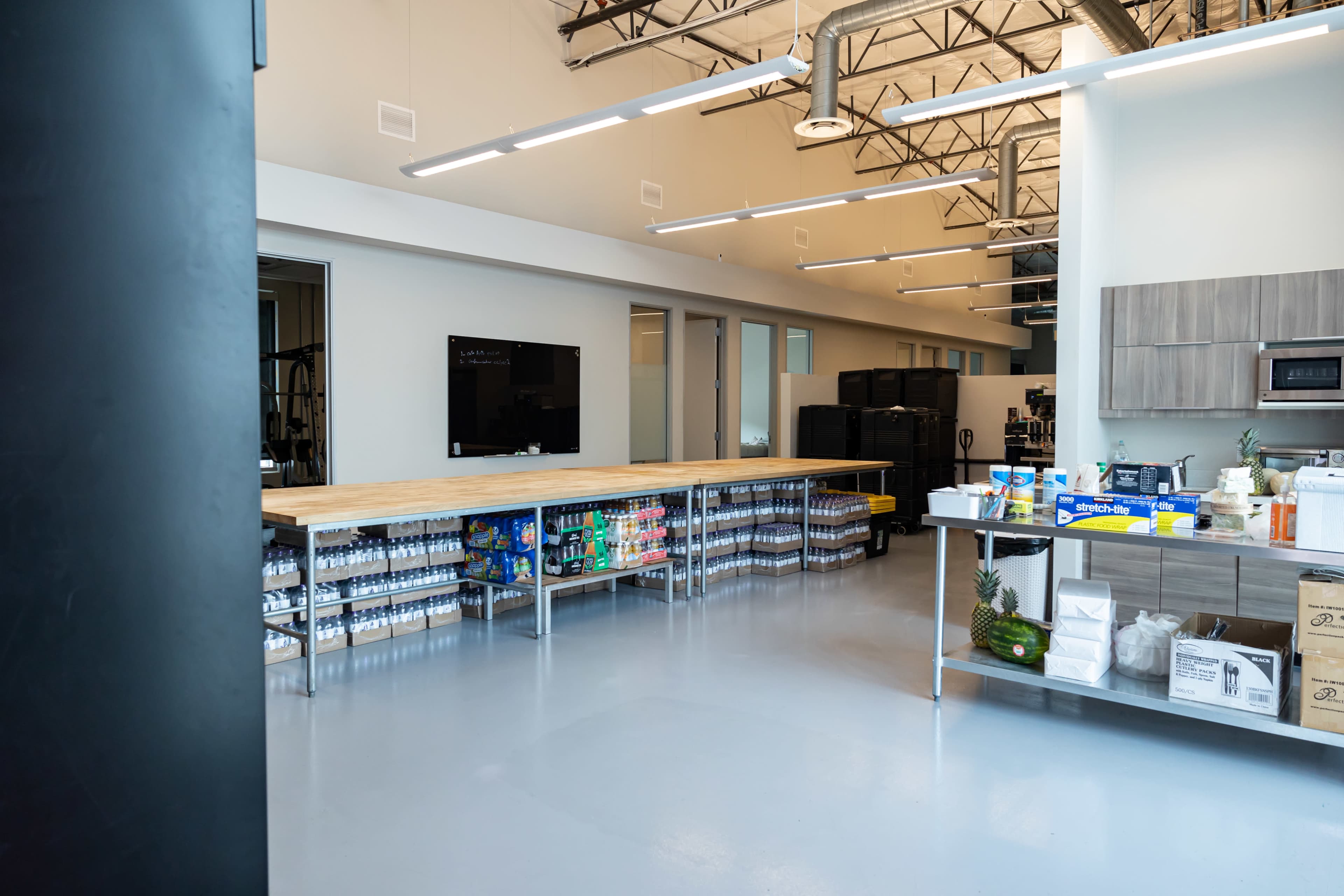 A spacious, modern kitchen area features a long wooden table, a sleek countertop with various supplies, and a television mounted on the wall.