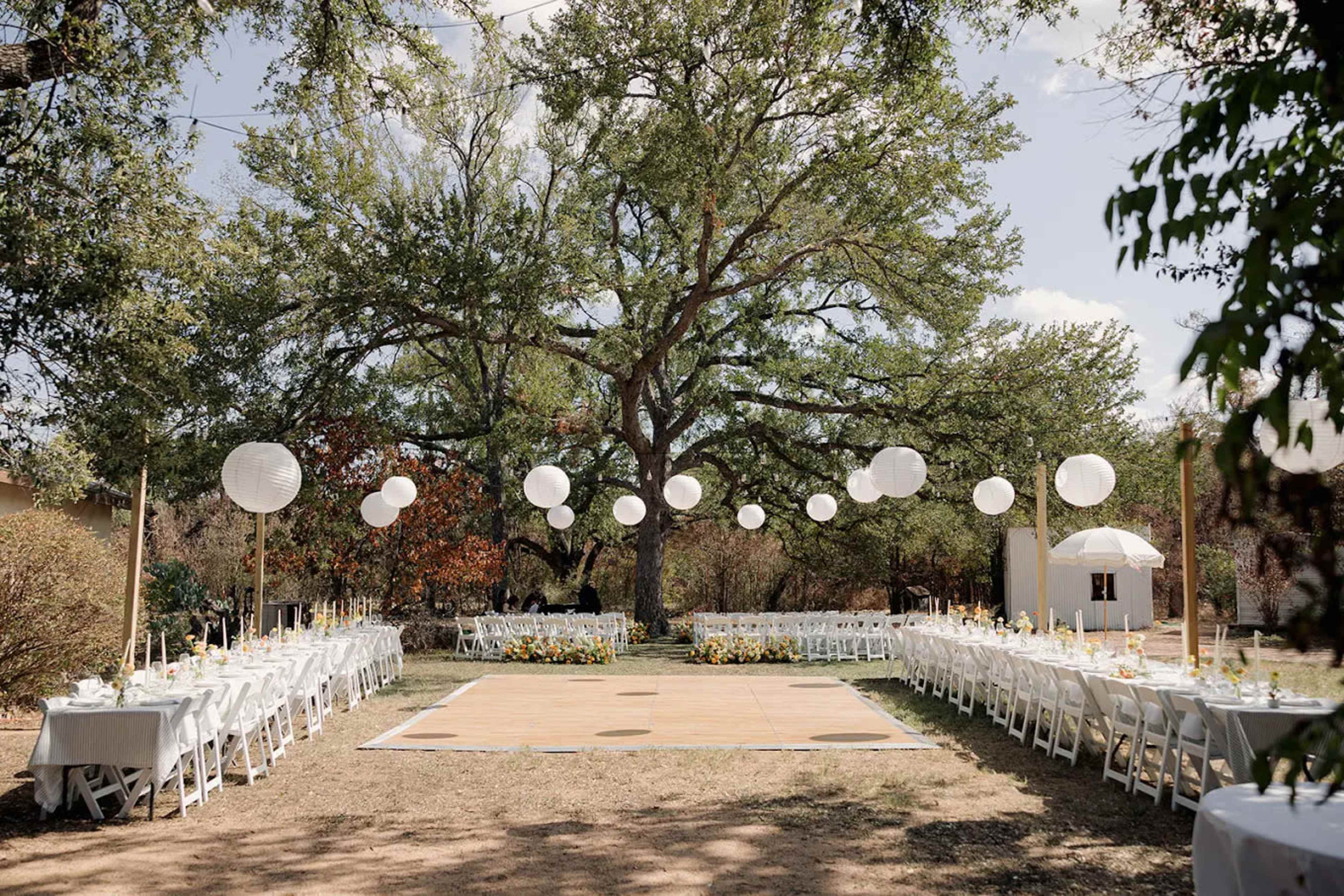 A decorated outdoor wedding venue features white tables arranged in two rows under a large tree, with hanging lanterns and a wooden dance floor at the center.