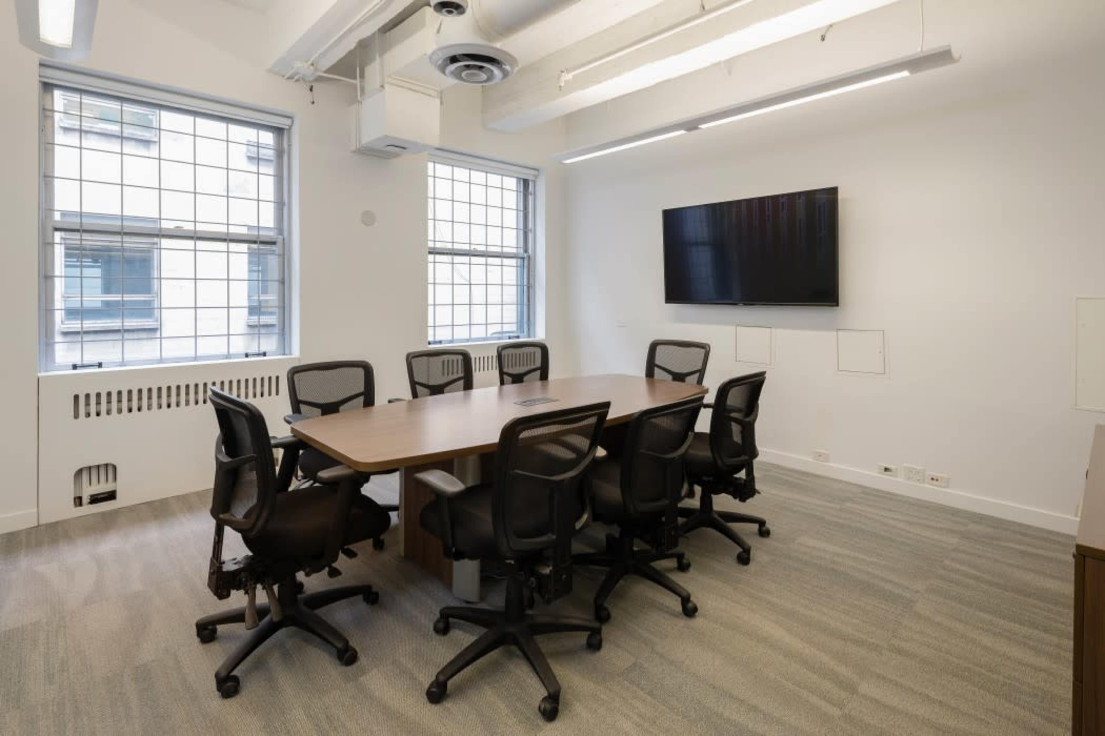 A modern conference room with a large table surrounded by black office chairs, featuring two windows and a wall-mounted television.