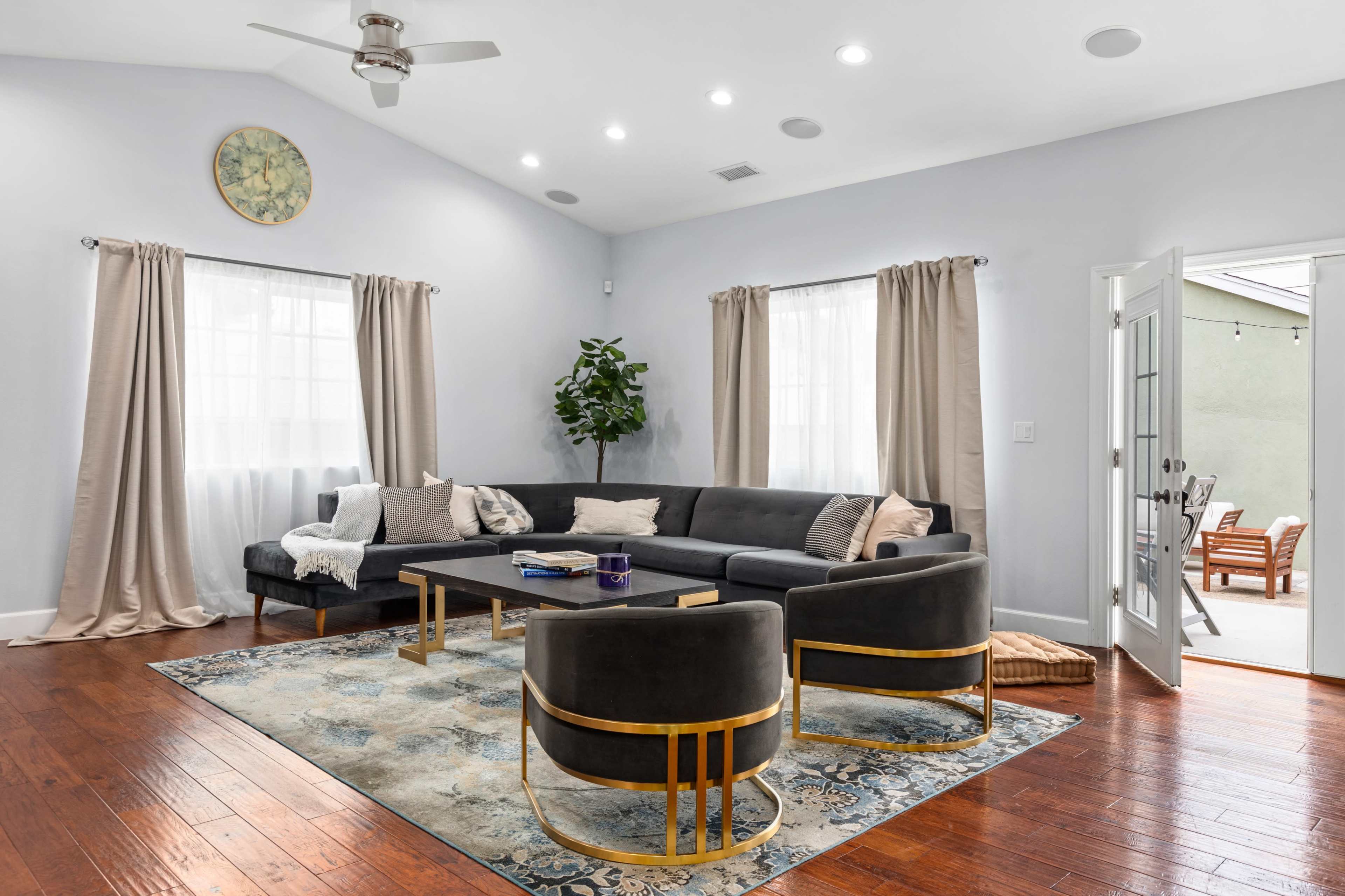 A modern living room features a large sectional sofa, two gold-accented chairs, a coffee table, and large windows with light curtains, all set on hardwood flooring.