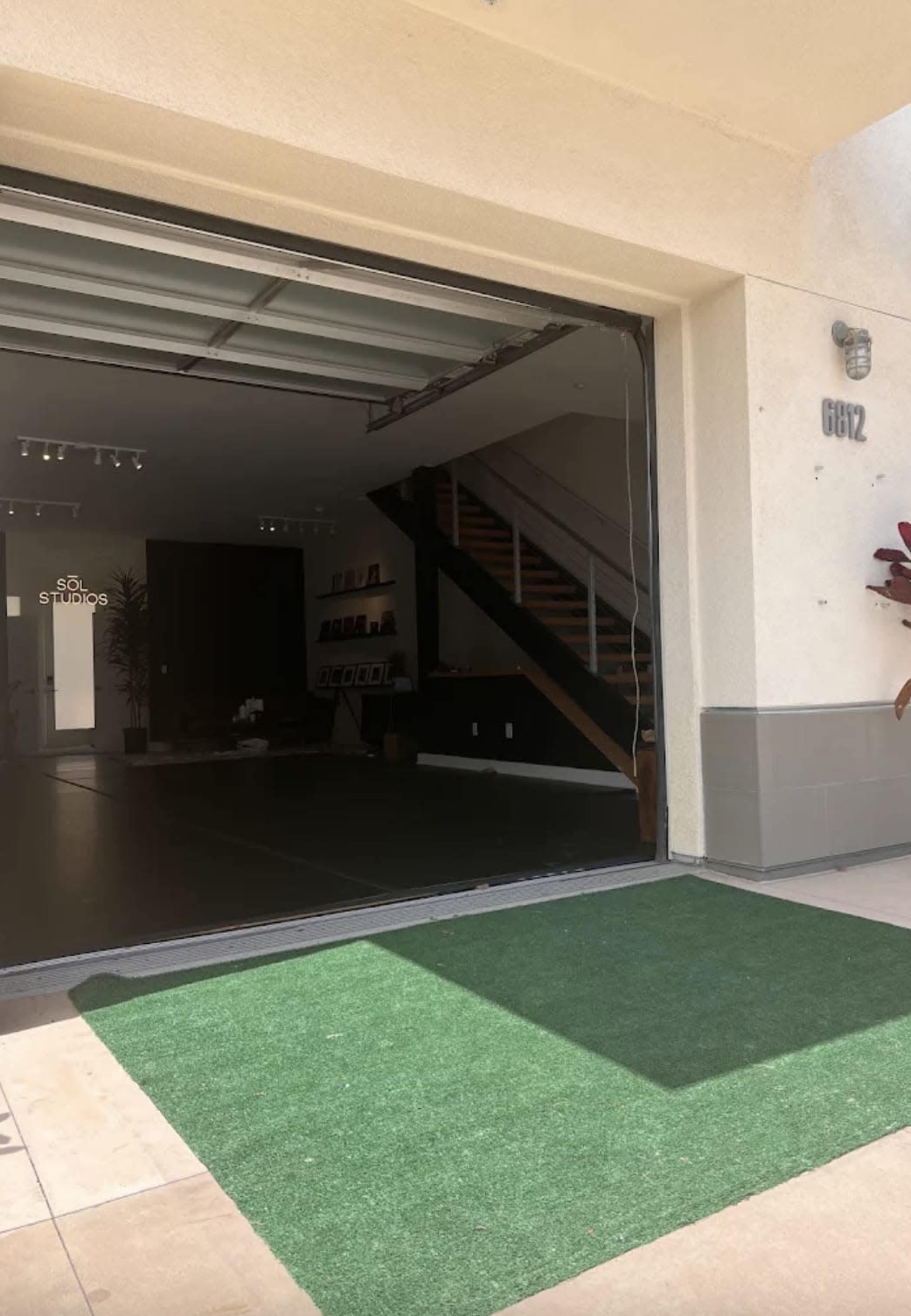 The image shows the entrance of a modern studio space with a large open garage door, revealing a staircase and interior decor.