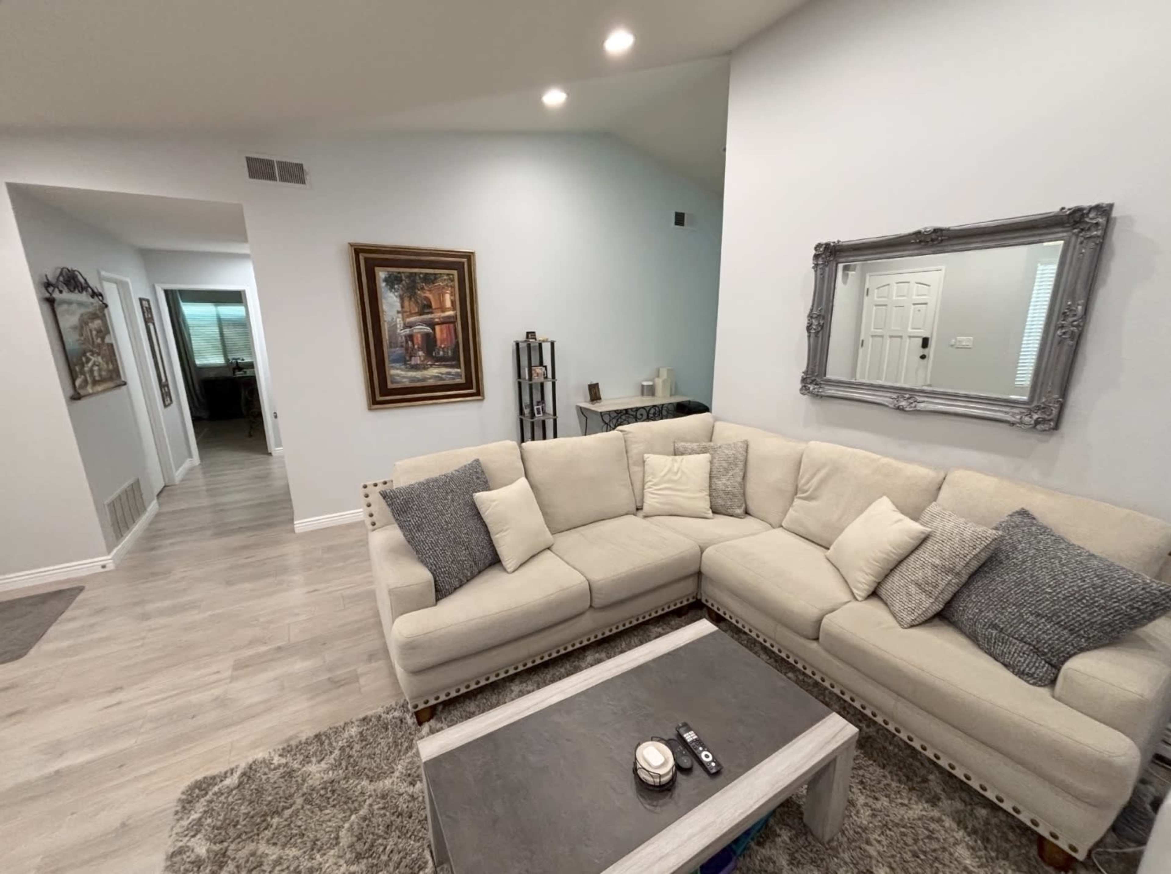 A light-colored sectional sofa with decorative pillows is positioned in a bright living room, with a coffee table in front and a hallway visible in the background.