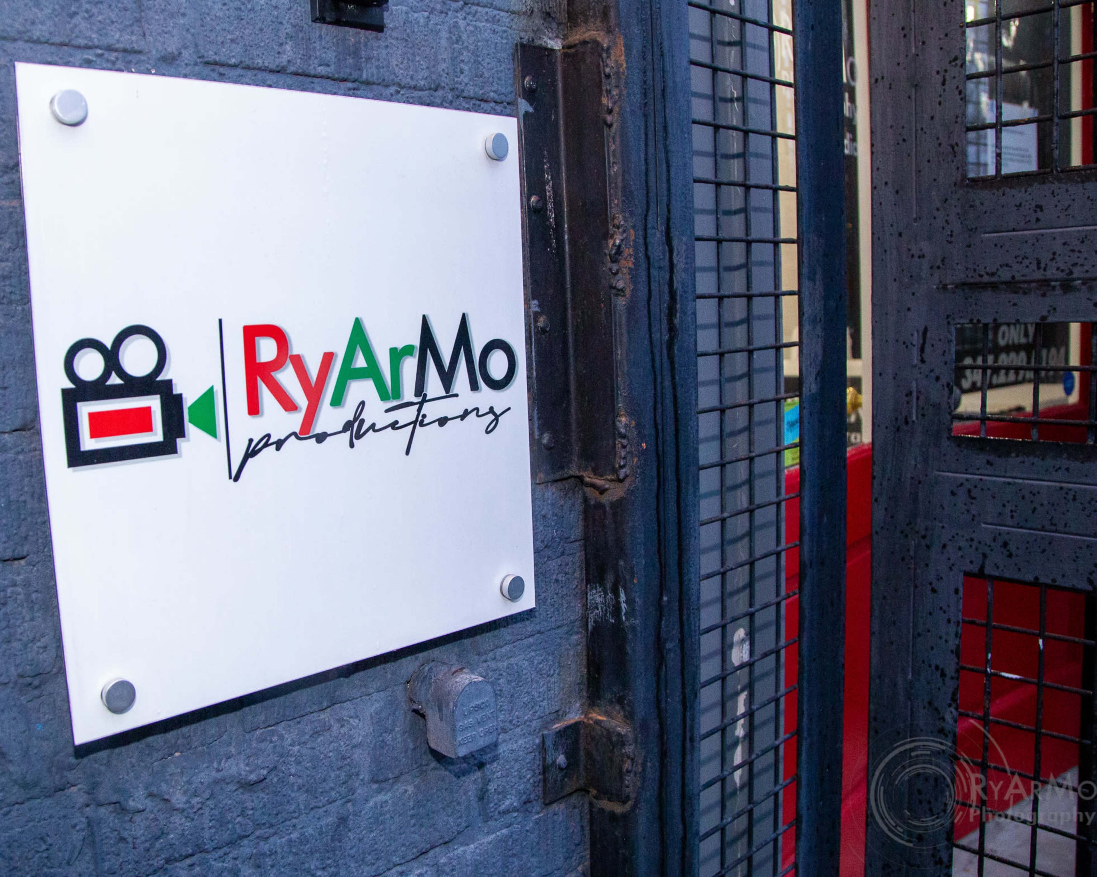 A sign for "RyArMo Productions" is displayed on a gray brick wall beside a metal gate.