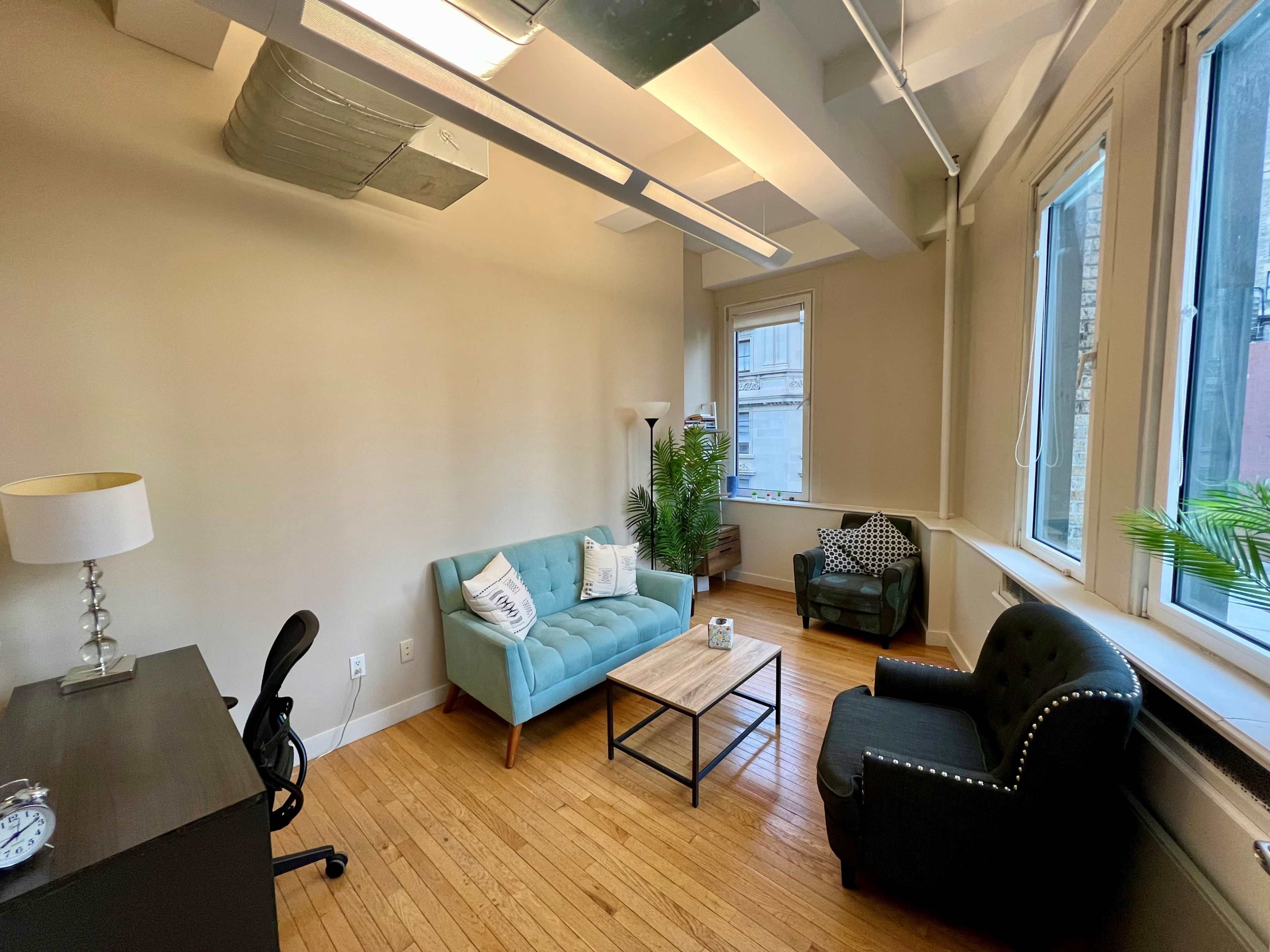 A bright office space furnished with a blue couch, a black armchair, a small coffee table, and a desk against the wall.