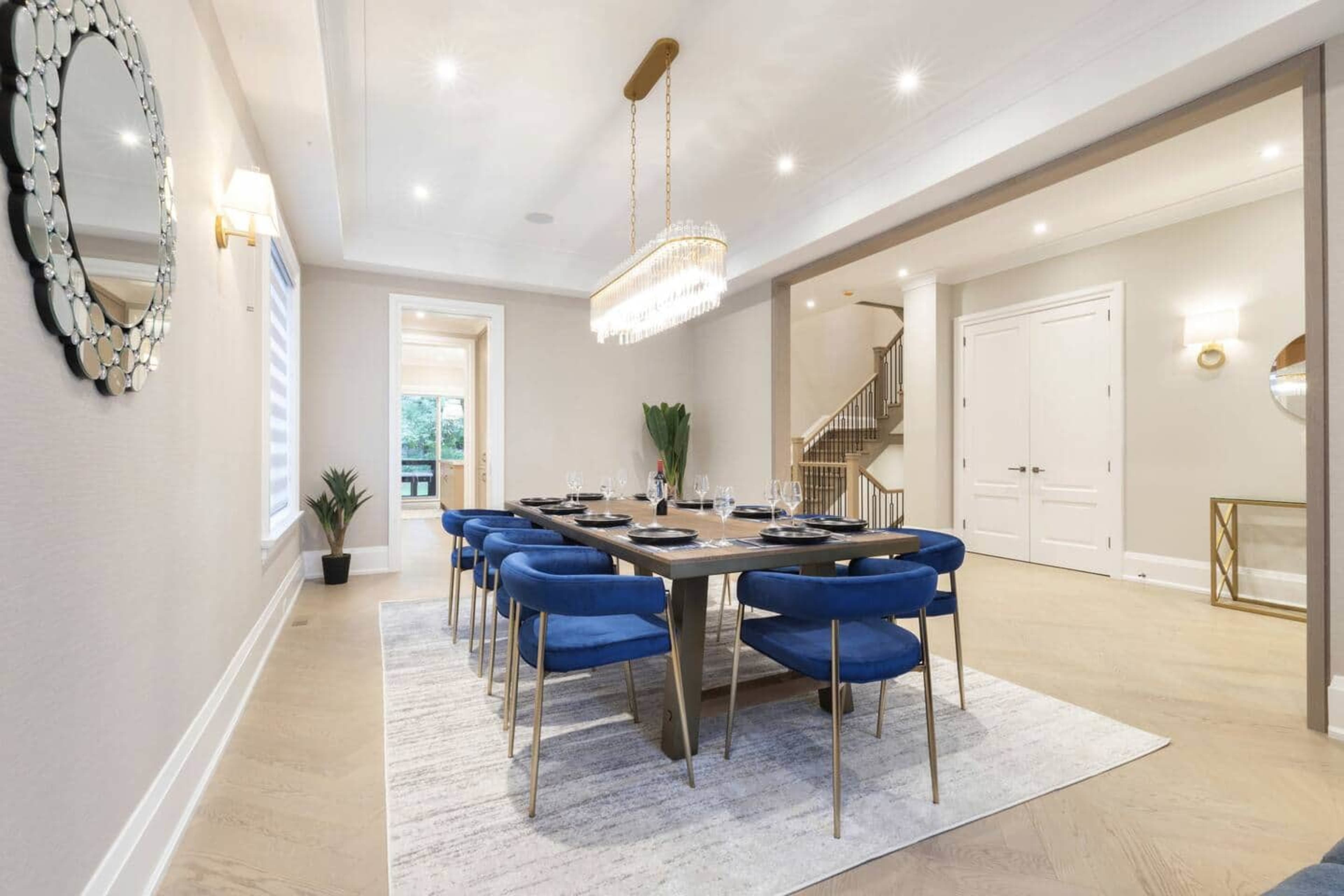 A spacious dining room features a large wooden table surrounded by blue chairs, a chandelier overhead, and stylish decor, including a mirror and potted plants.