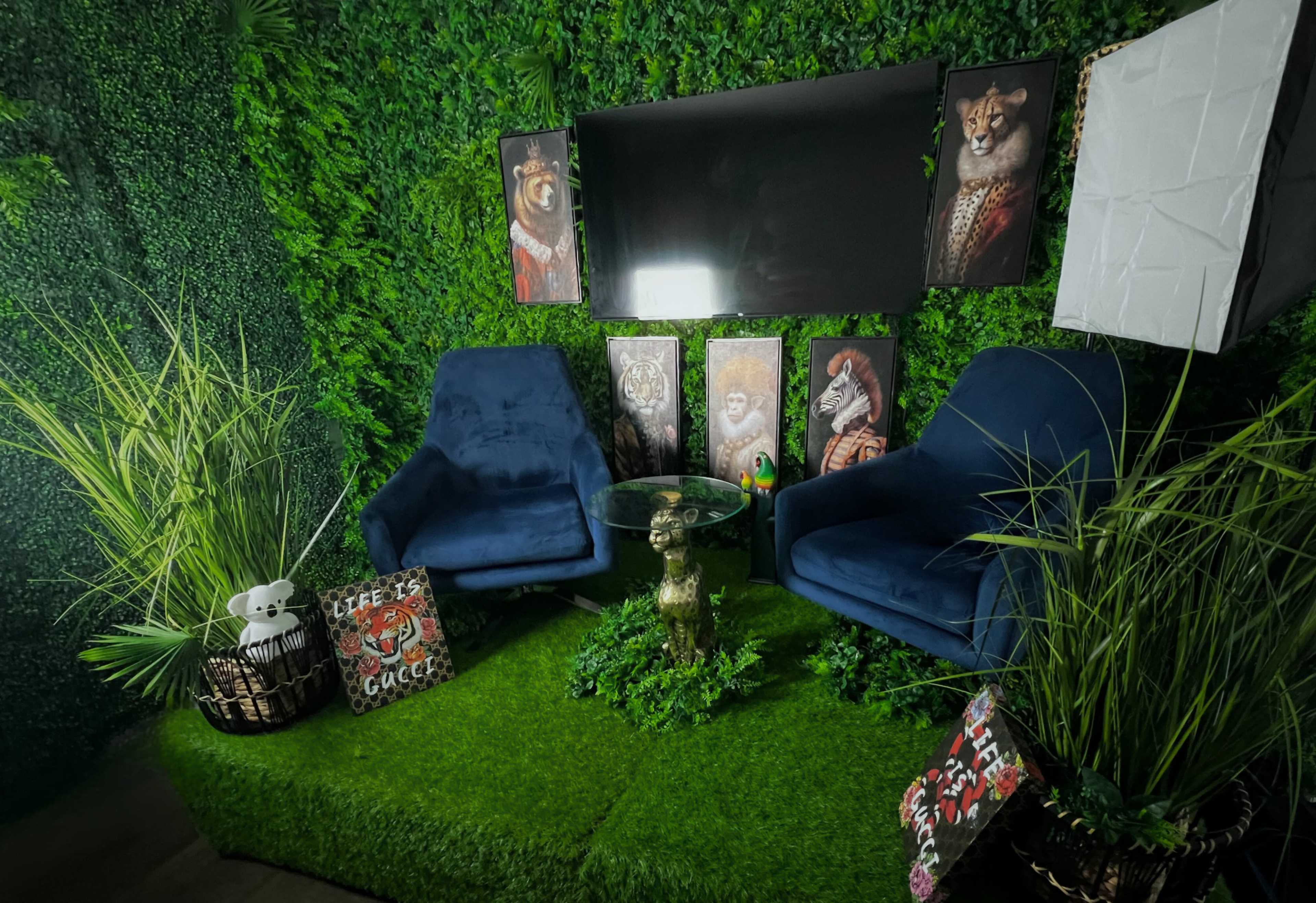 The image shows a cozy seating area with two blue chairs, a glass table, decorative animal art on the walls, and a lush green plant backdrop.