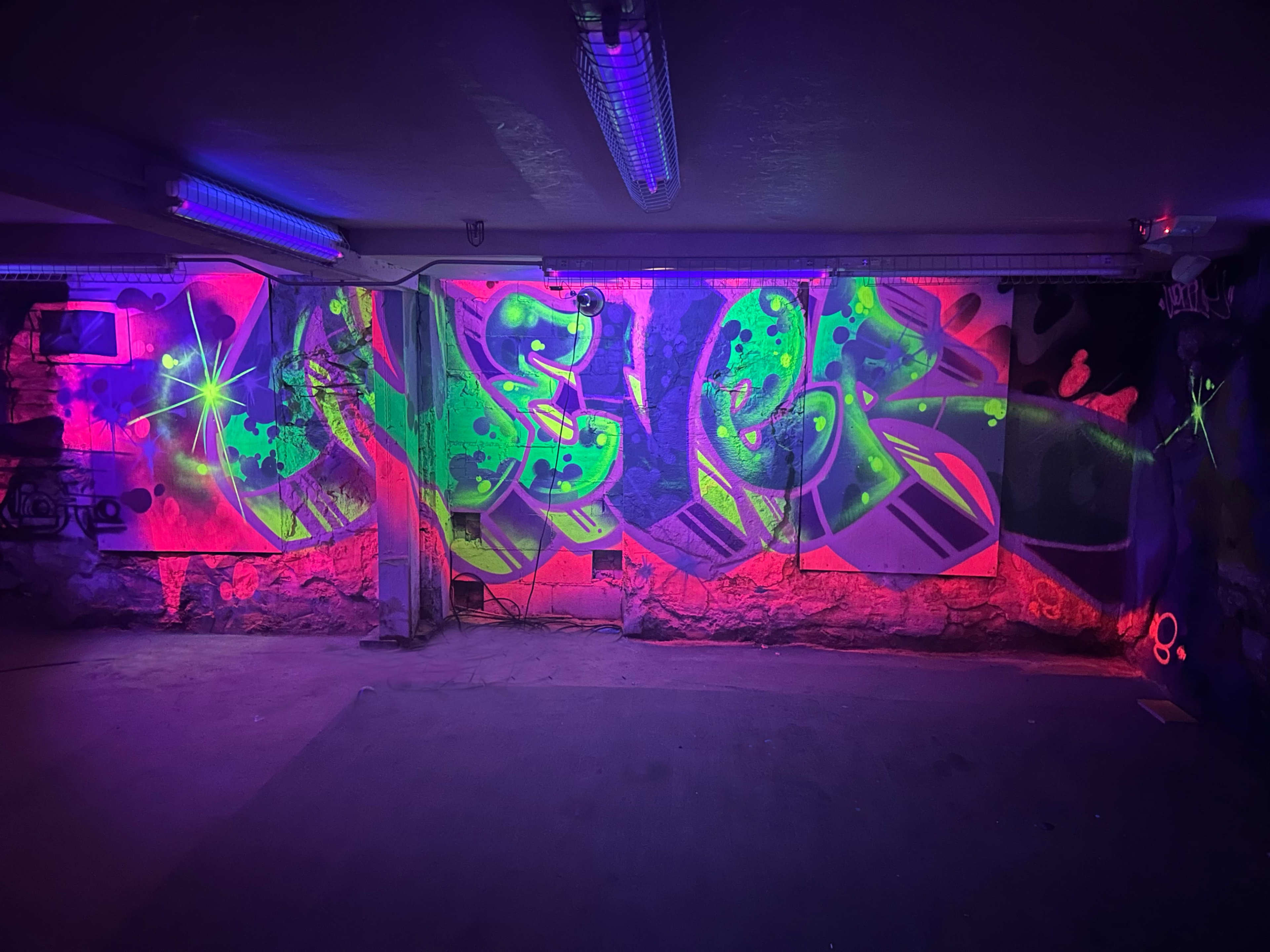 A vibrant mural of graffiti on a wall, illuminated by purple and green lighting in a dimly lit underground space.