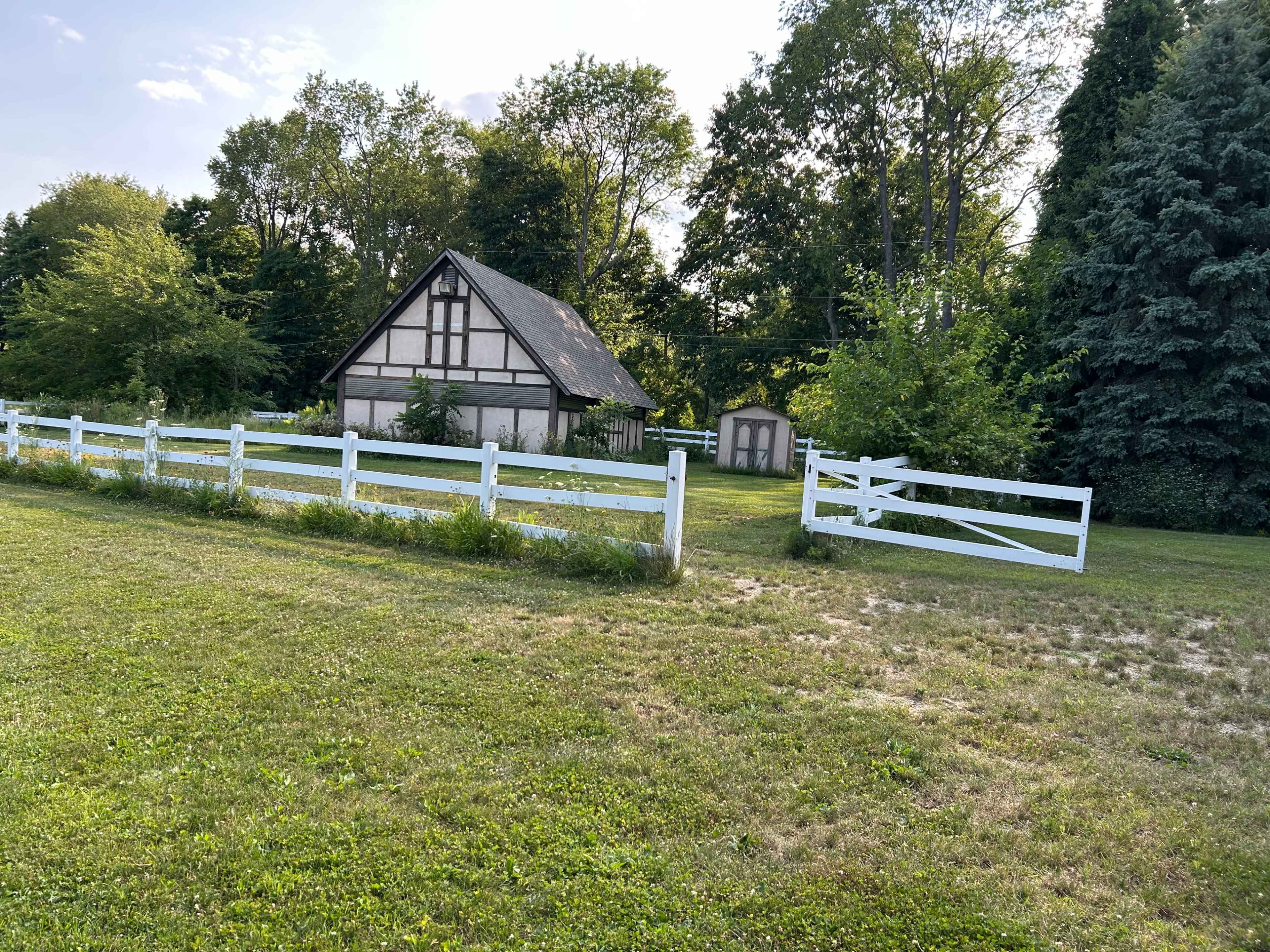 A large, rustic house with a sloped roof is surrounded by a white wooden fence and landscaped greenery.