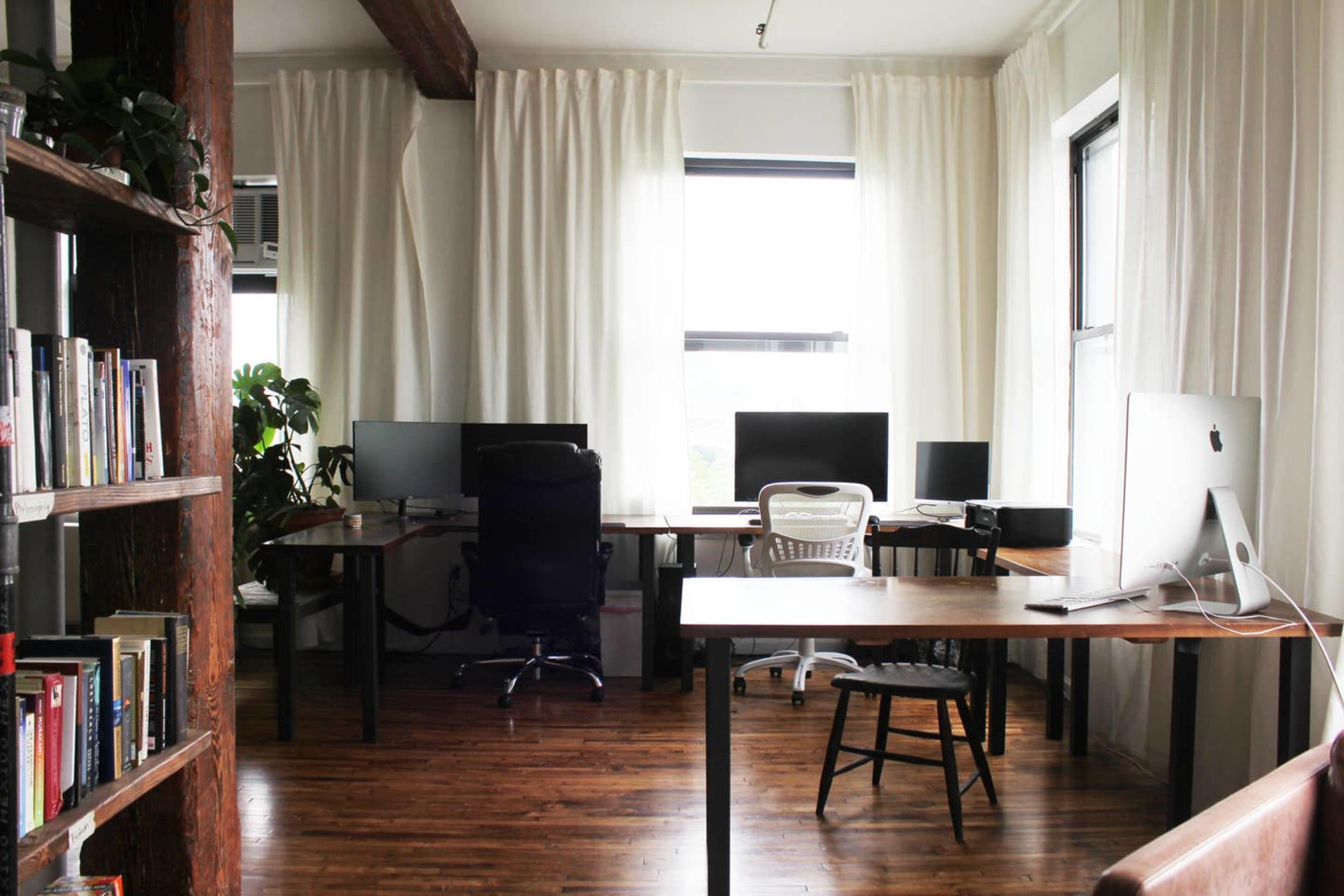 A workspace with two desks, multiple computer monitors, a chair, and bookshelves, all set in a room with large windows and curtains.