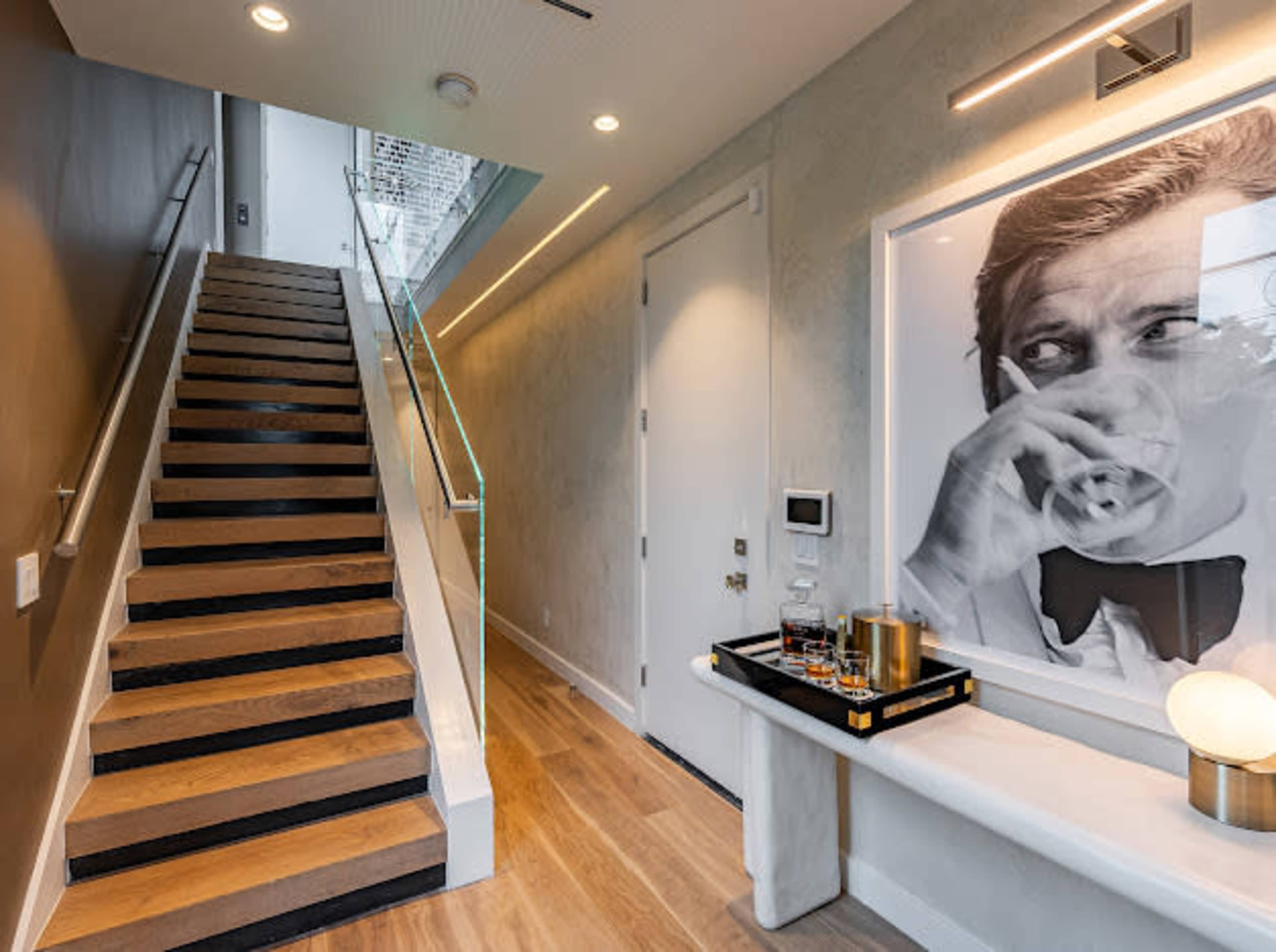 The image shows a modern entryway with a staircase leading upward, a large black-and-white portrait on the wall, and a side table featuring a decorative tray with glassware and a lamp.
