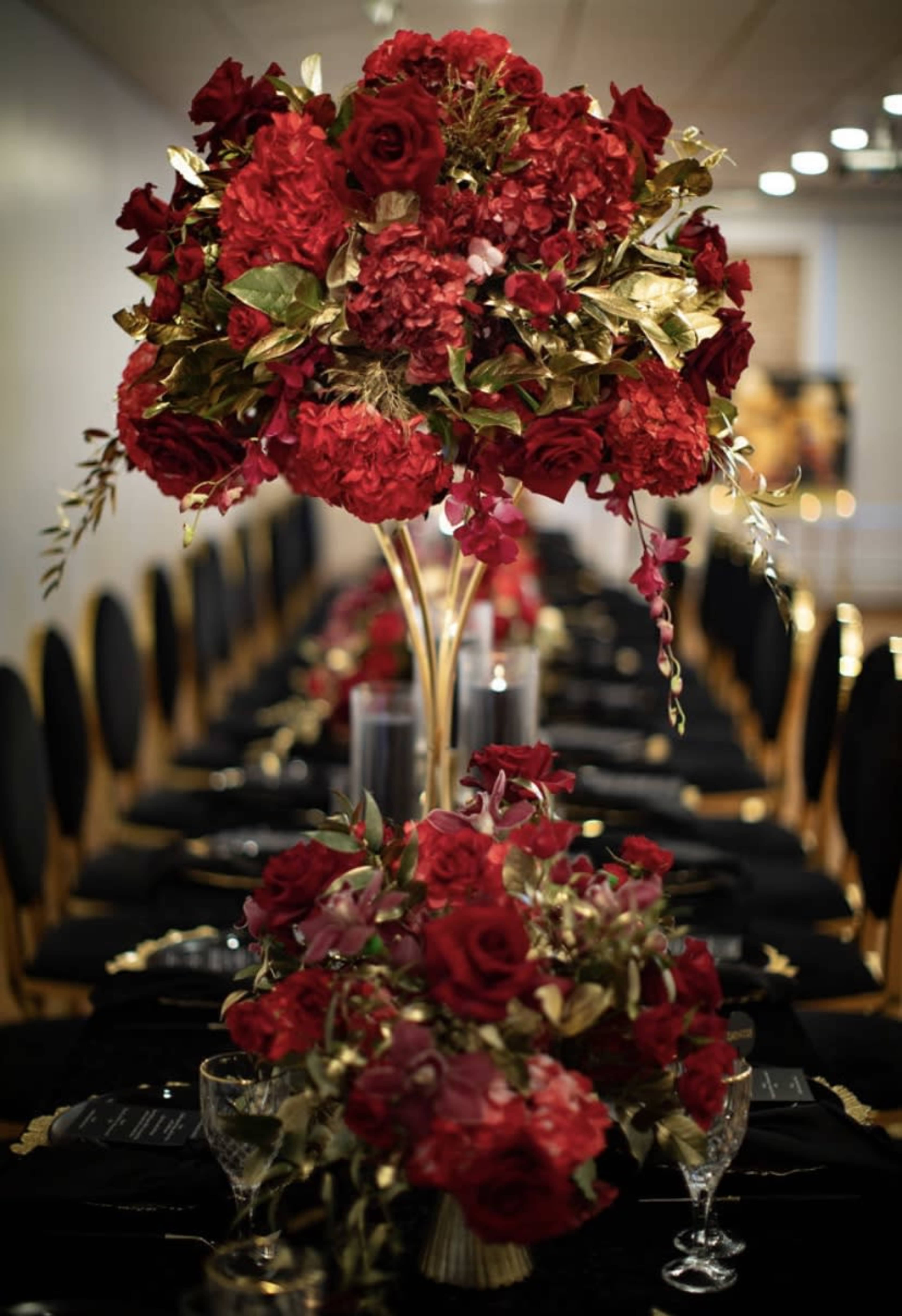 A dining table is elegantly set with tall floral centerpieces made of red roses and hydrangeas, surrounded by smaller arrangements and black table settings.