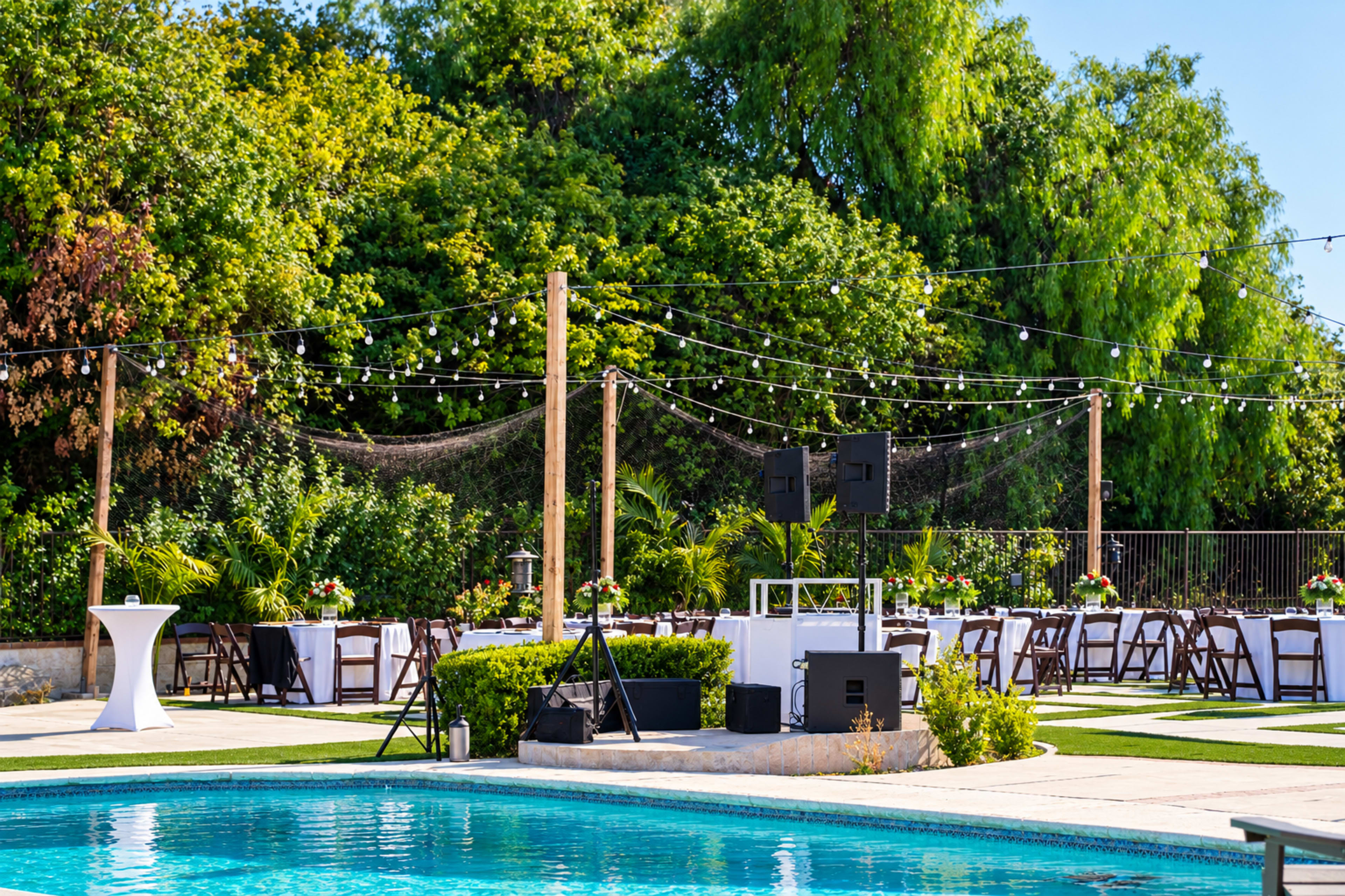 A landscaped outdoor area features a pool surrounded by tables set for an event, with string lights overhead and greenery in the background.