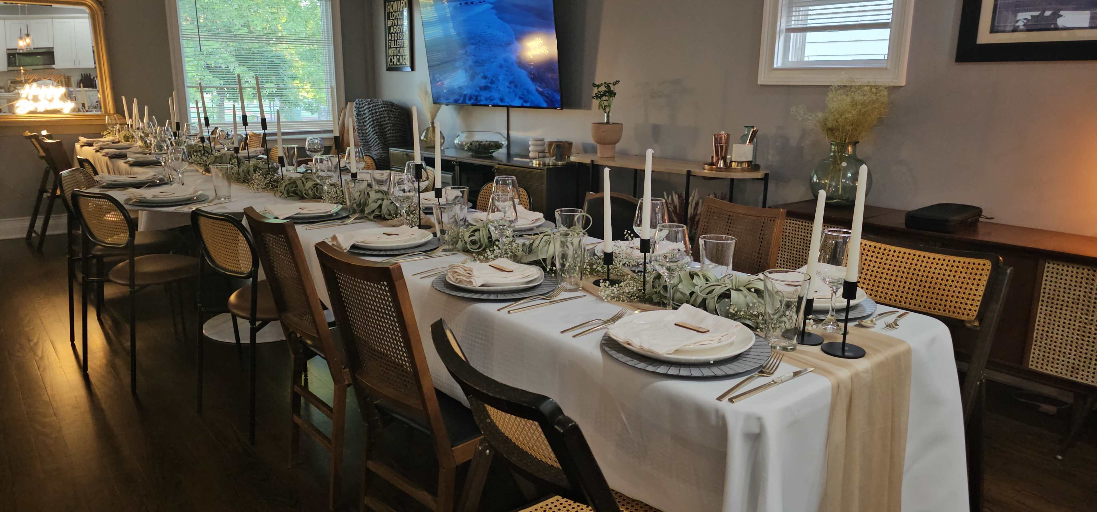 A long dining table is set for a formal meal, adorned with white tablecloths, plates, glassware, and candles, in a well-lit room with decorative elements.