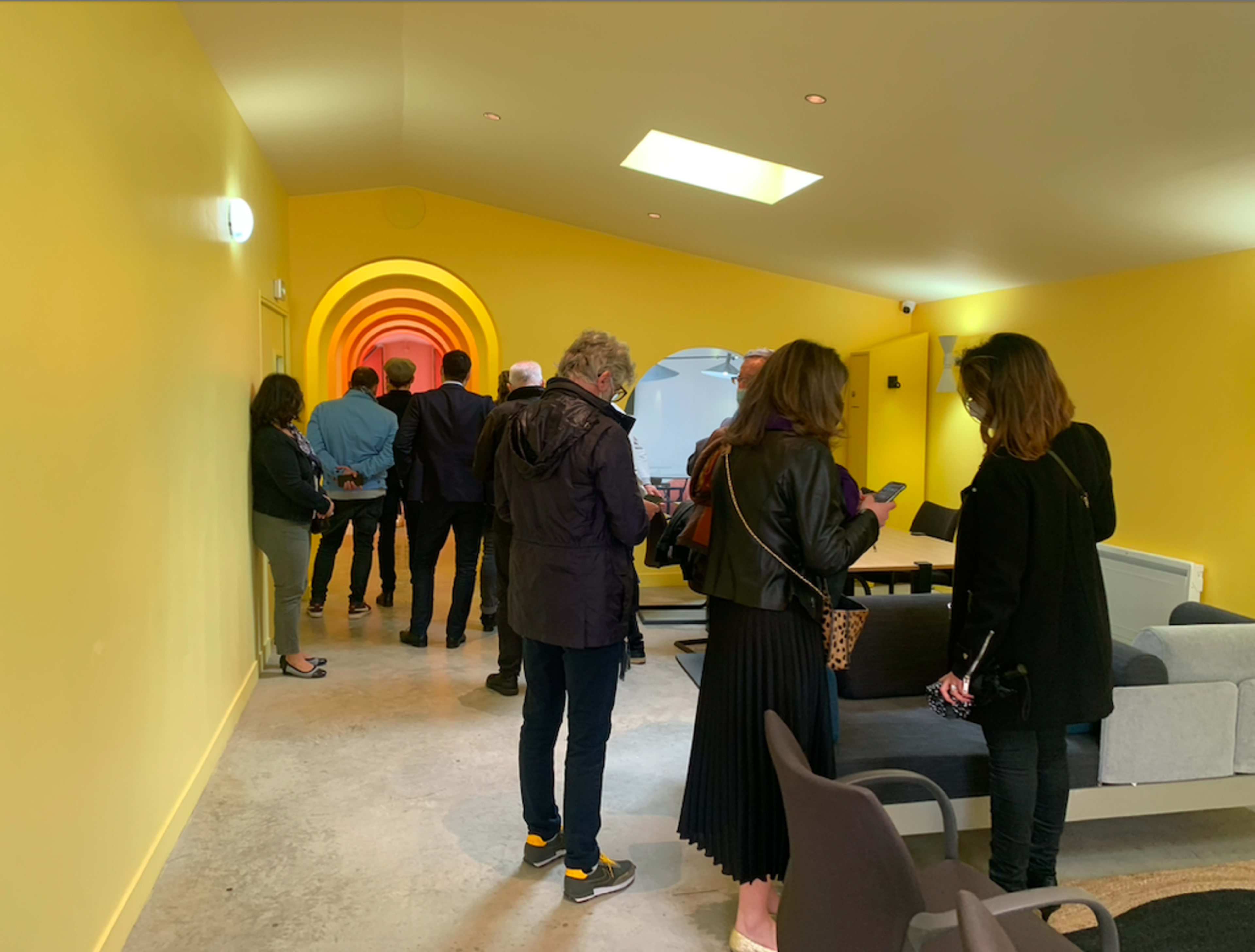 A group of people stands in a yellow hallway leading to a brightly lit archway.