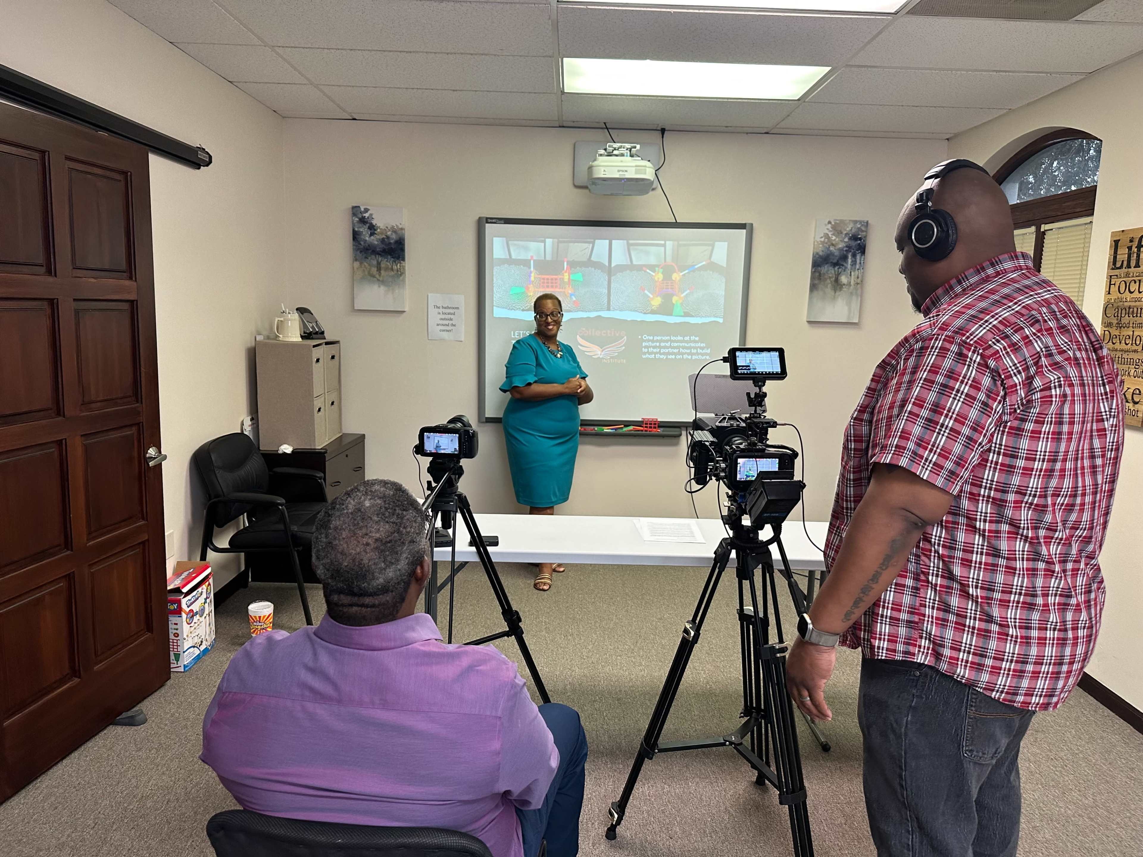 A woman stands in front of a projector screen while two men operate cameras in a classroom setting.