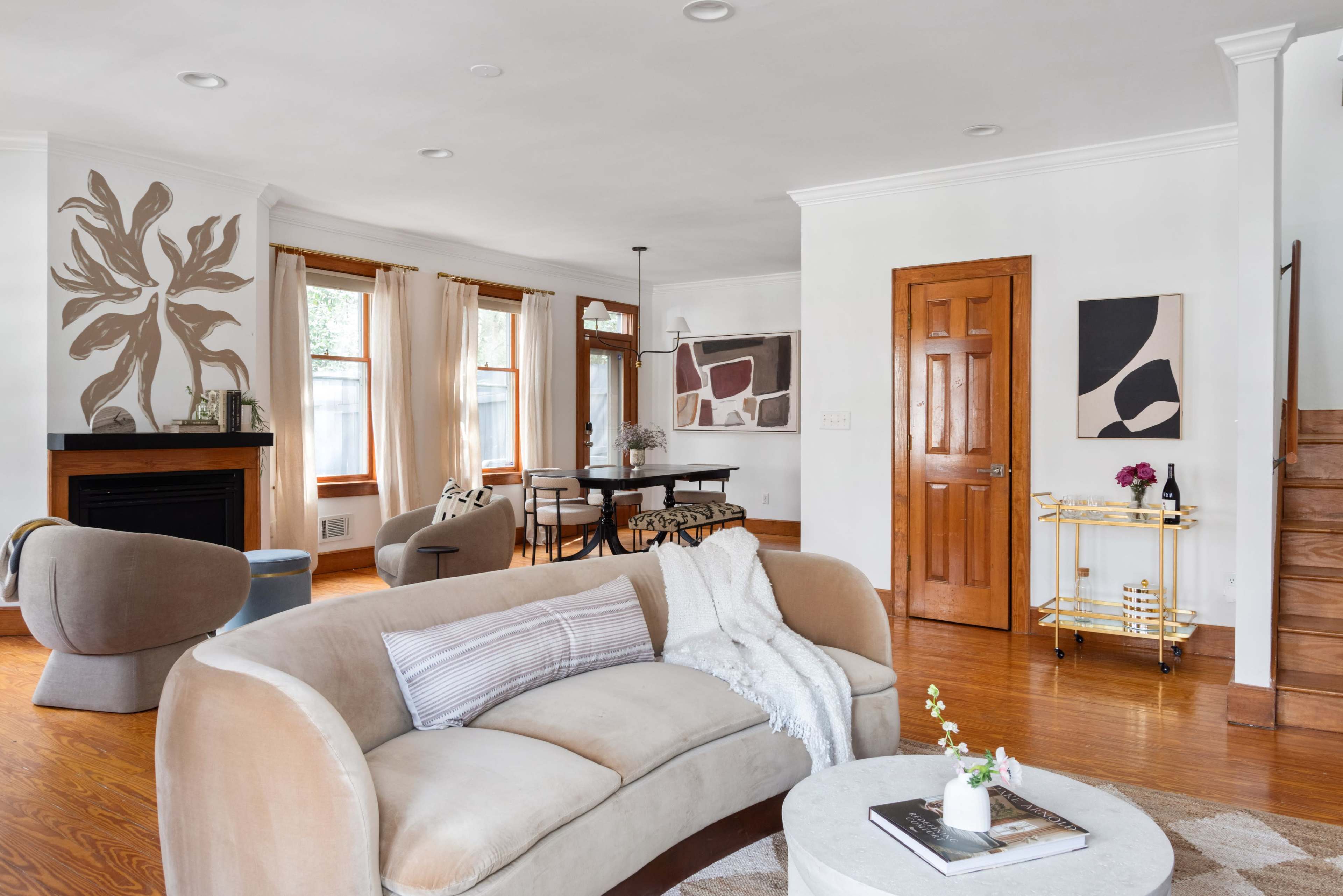 The living room features a soft sectional sofa, a round coffee table, and a dining area with a wooden table, all set within a well-lit space that includes decorative artwork and a staircase.