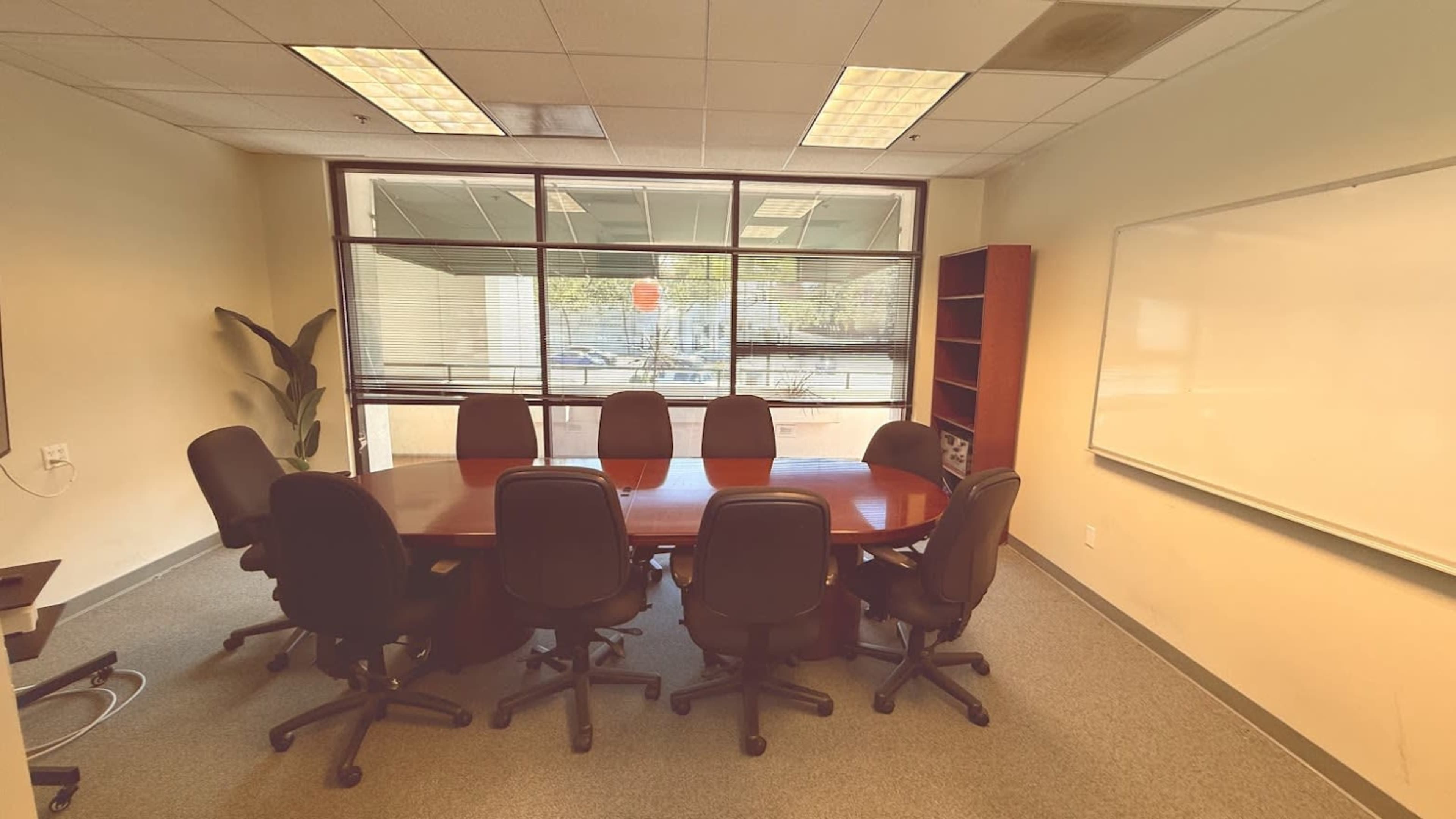 A conference room features a large oval table surrounded by eight black chairs, with windows providing natural light and a whiteboard on one wall.