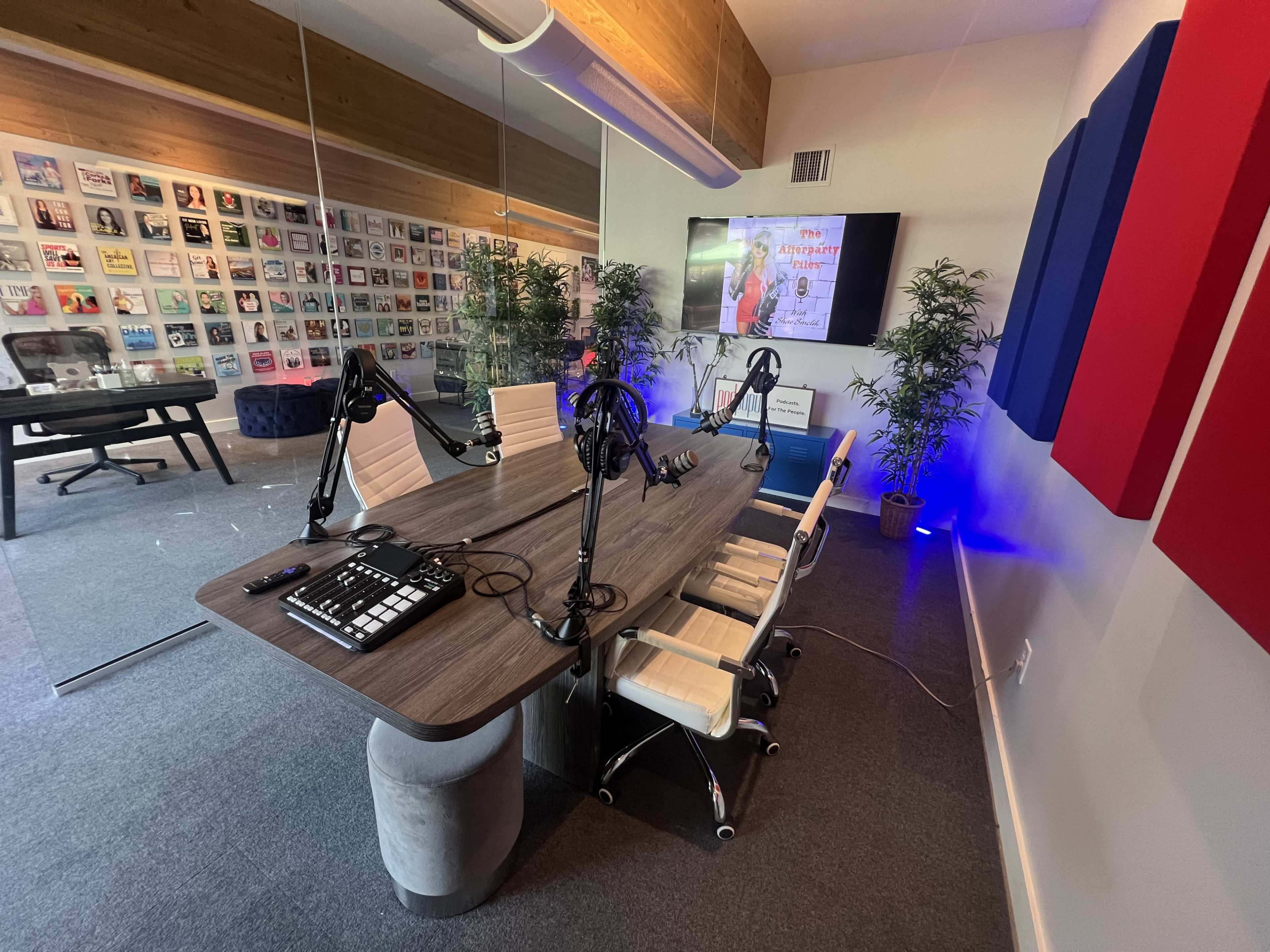 The image shows a modern podcasting studio with a large table equipped with microphones, headphones, and audio equipment, surrounded by potted plants and featuring a screen displaying content on one wall.