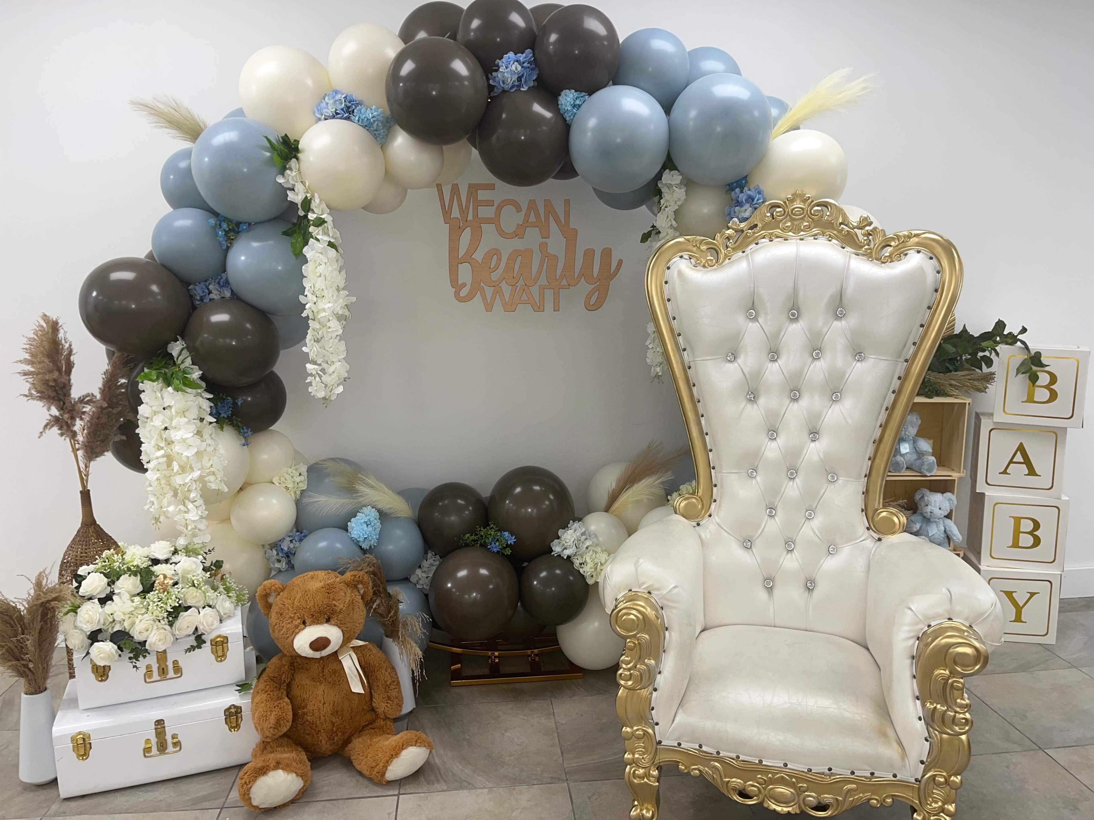 A decorative setup for a baby-themed event, including a balloon arch, a teddy bear, and a plush white throne chair.