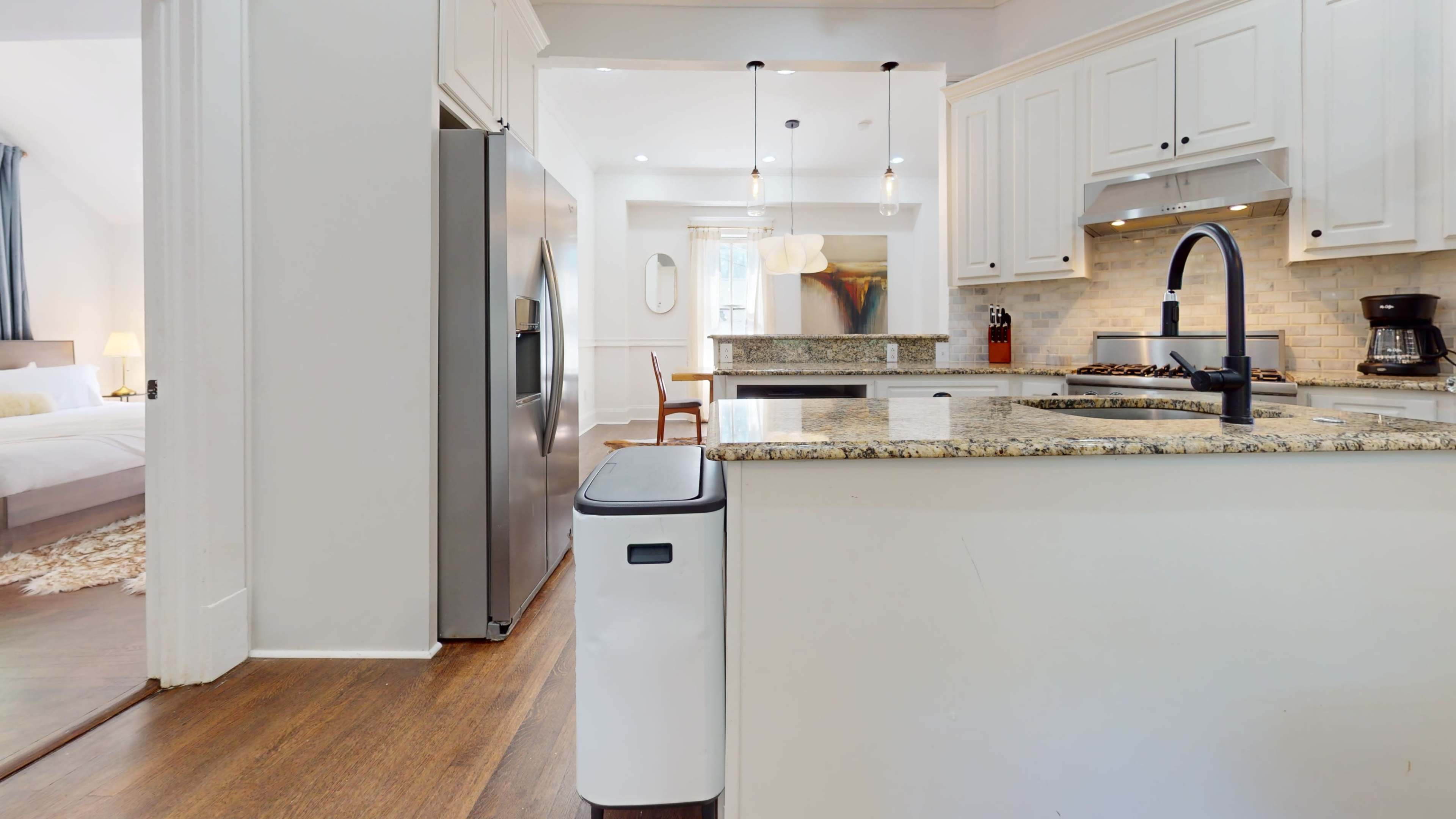 The image shows a modern kitchen with a stainless steel refrigerator, granite countertops, and a white waste bin beside the sink.