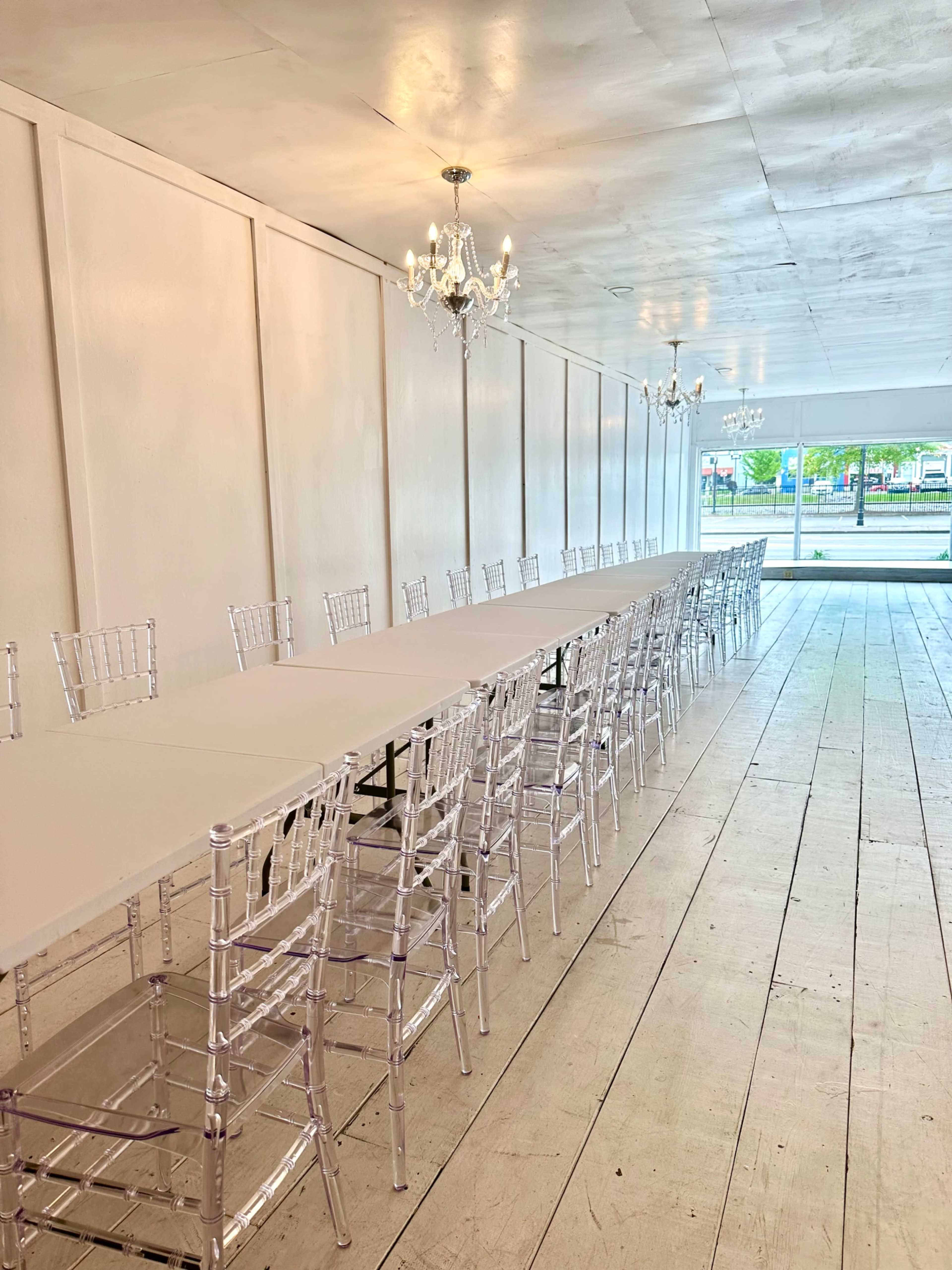 A long table is set up with transparent chairs in a well-lit room featuring white walls and a chandelier.