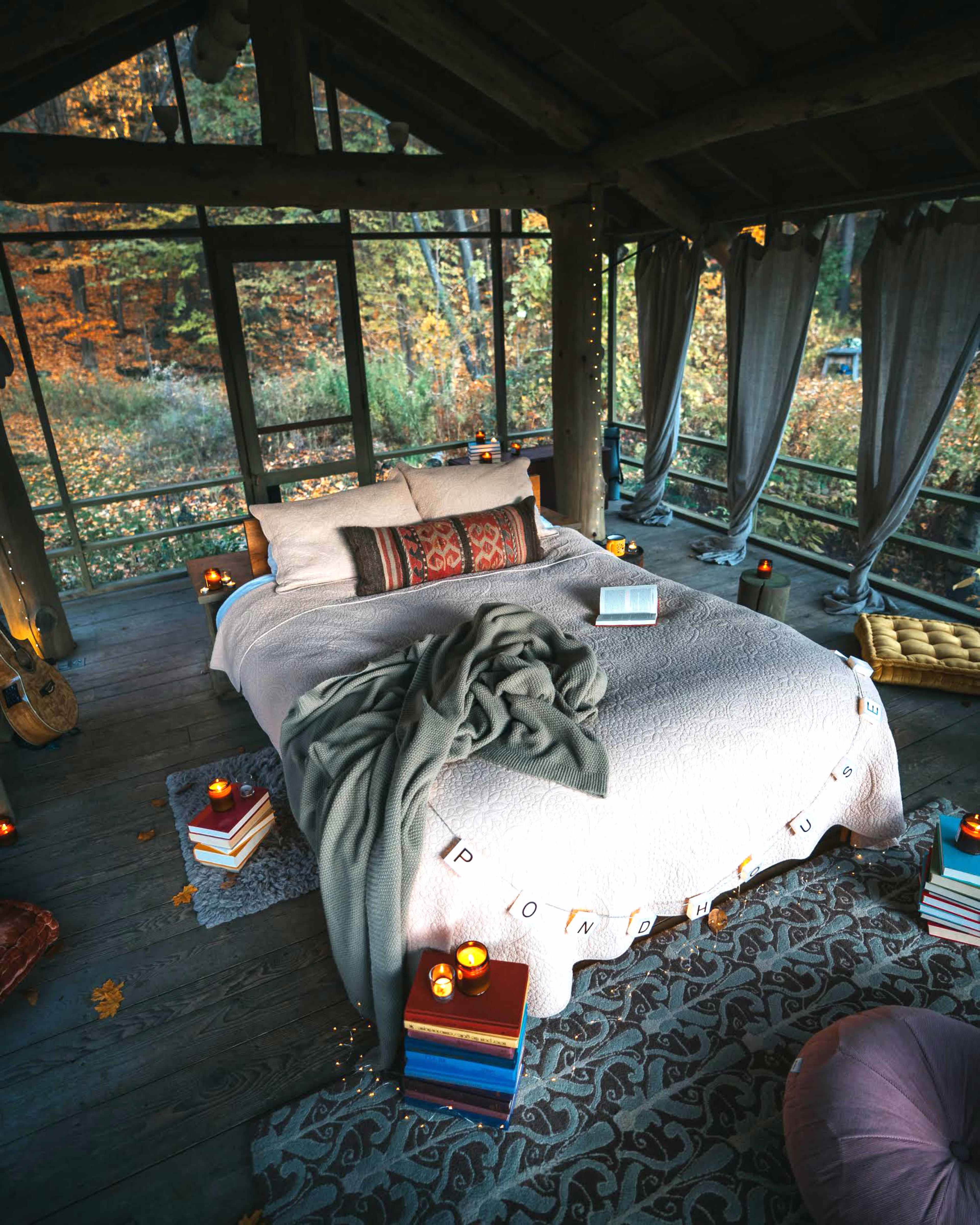 A cozy bedroom features a neatly made bed surrounded by candles, books, and soft furnishings, with large windows displaying autumn foliage outside.