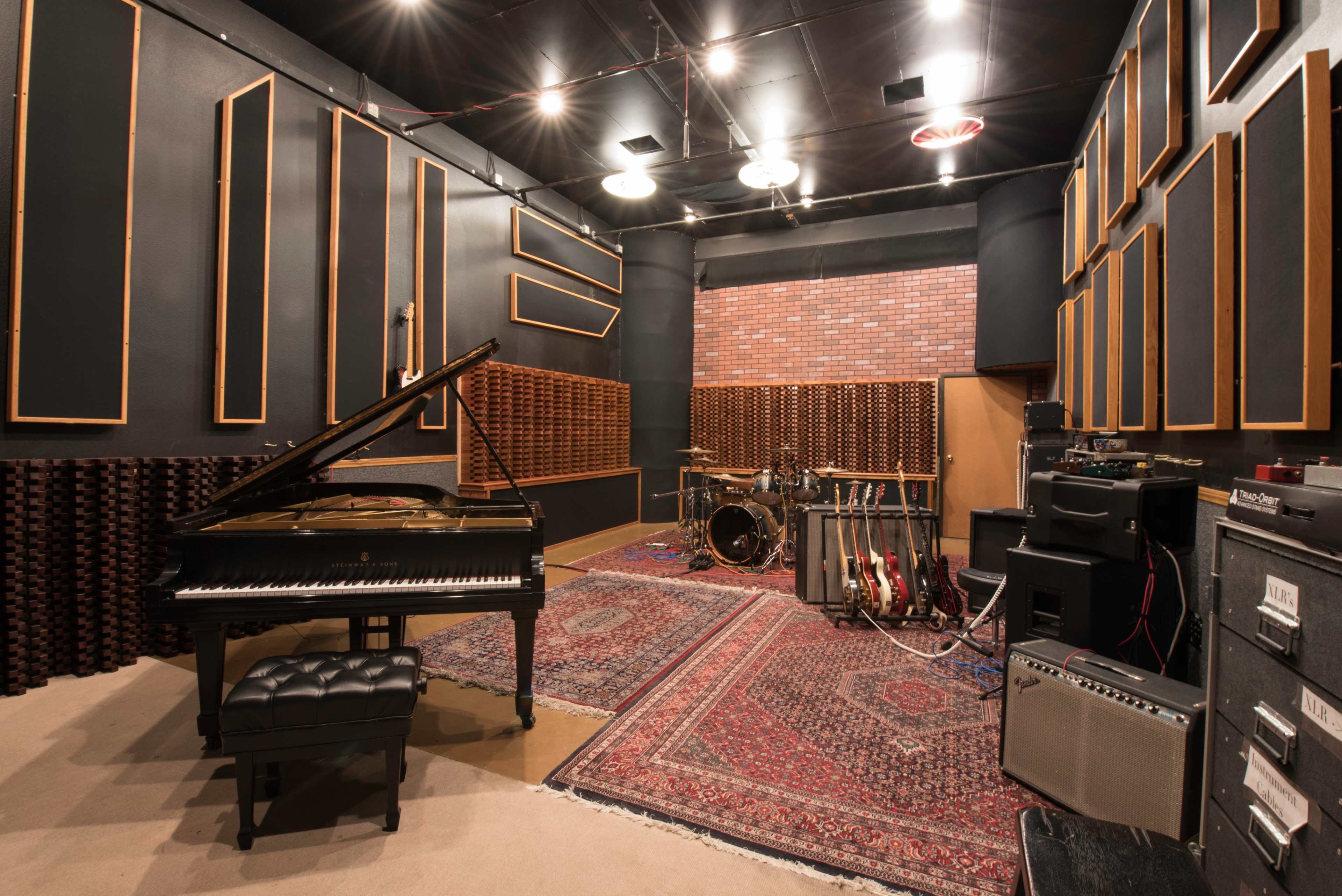 A music recording studio featuring a grand piano, drum set, electric guitars, and amplifiers arranged against soundproofed walls.