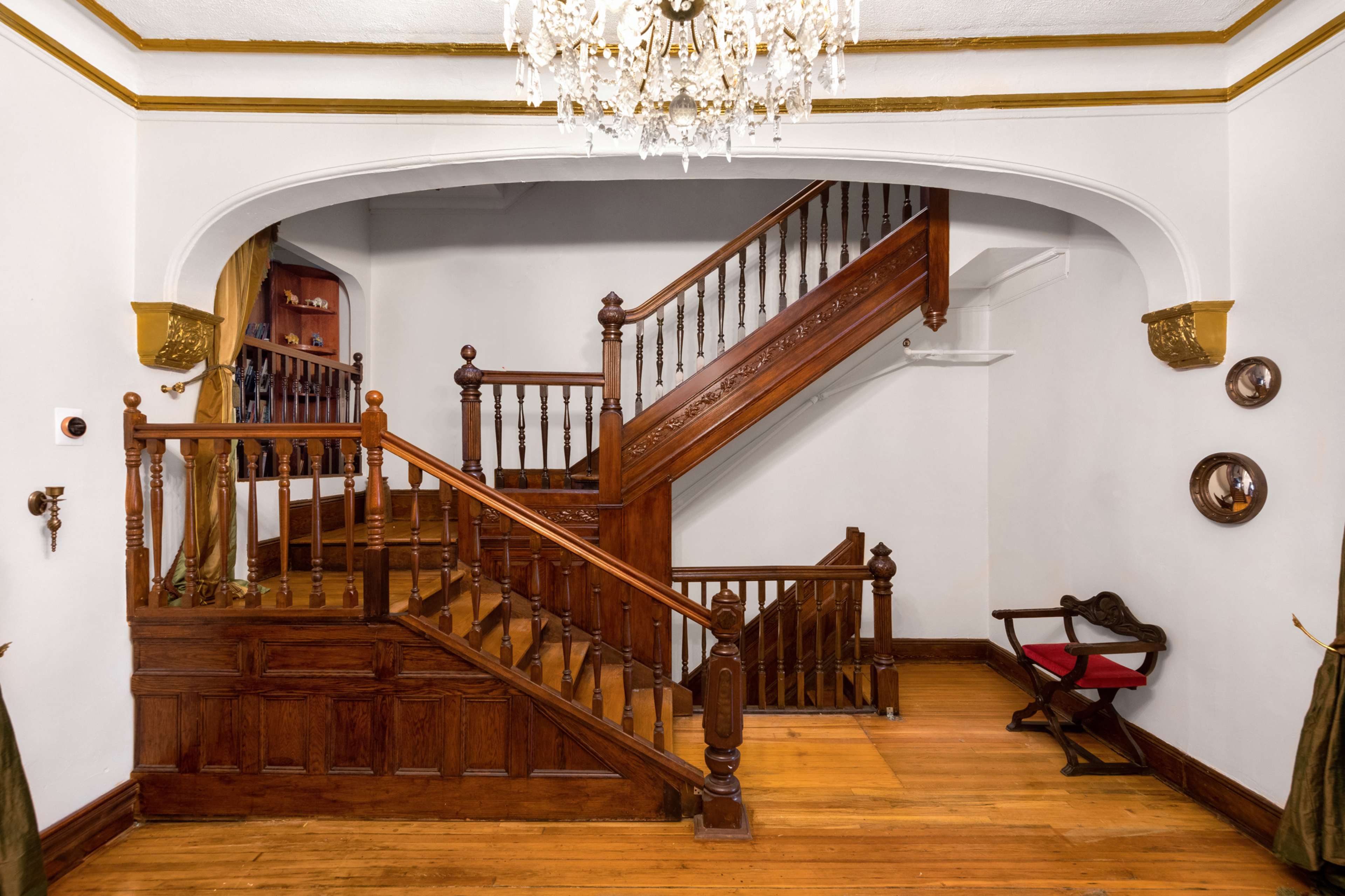 A wooden staircase with an ornate railing and a chandelier overhead in a well-lit foyer.