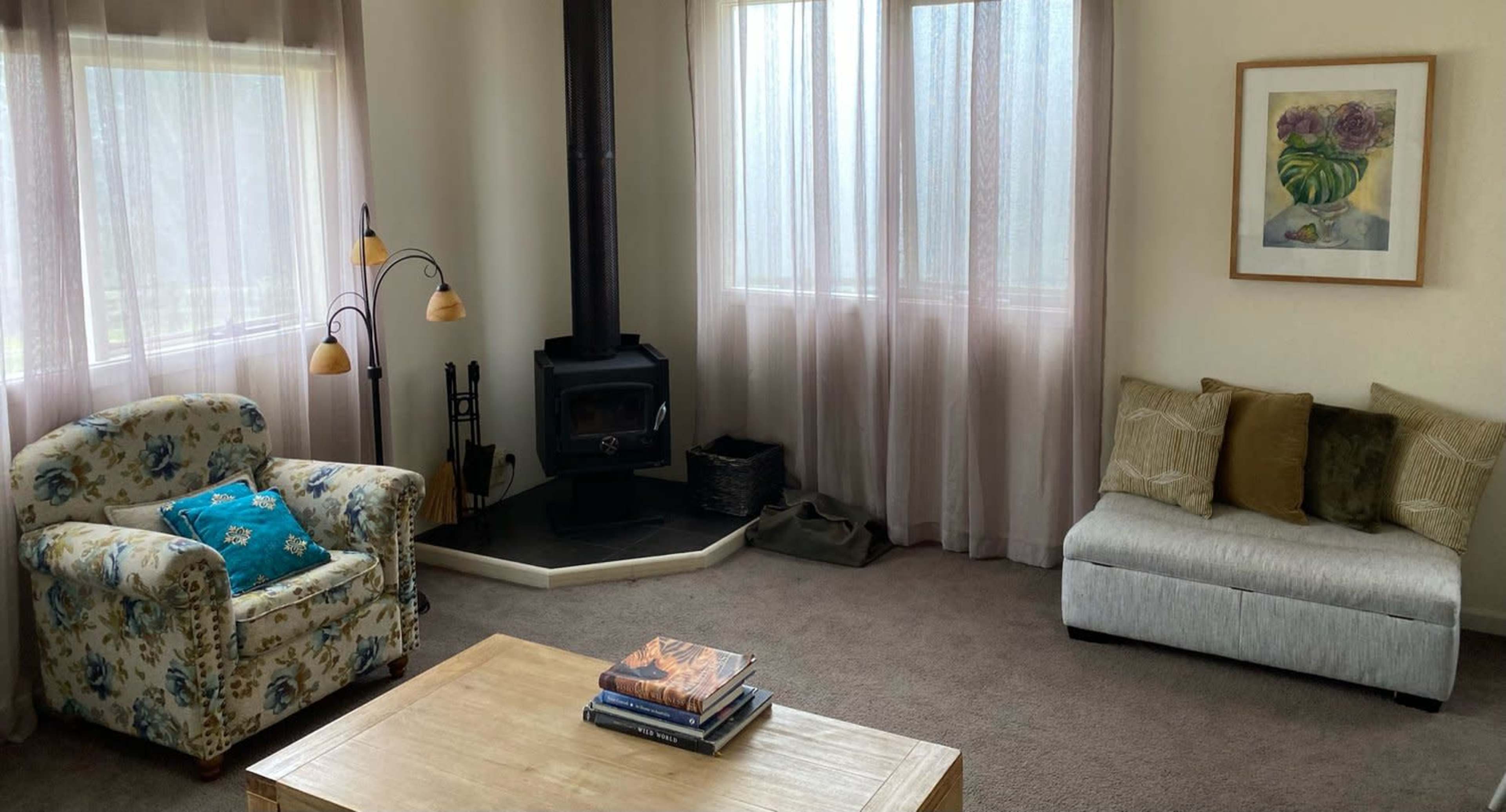 The image shows a living room with a floral-patterned armchair, a gray couch, a wooden coffee table, a wood stove, and large windows covered with sheer curtains.